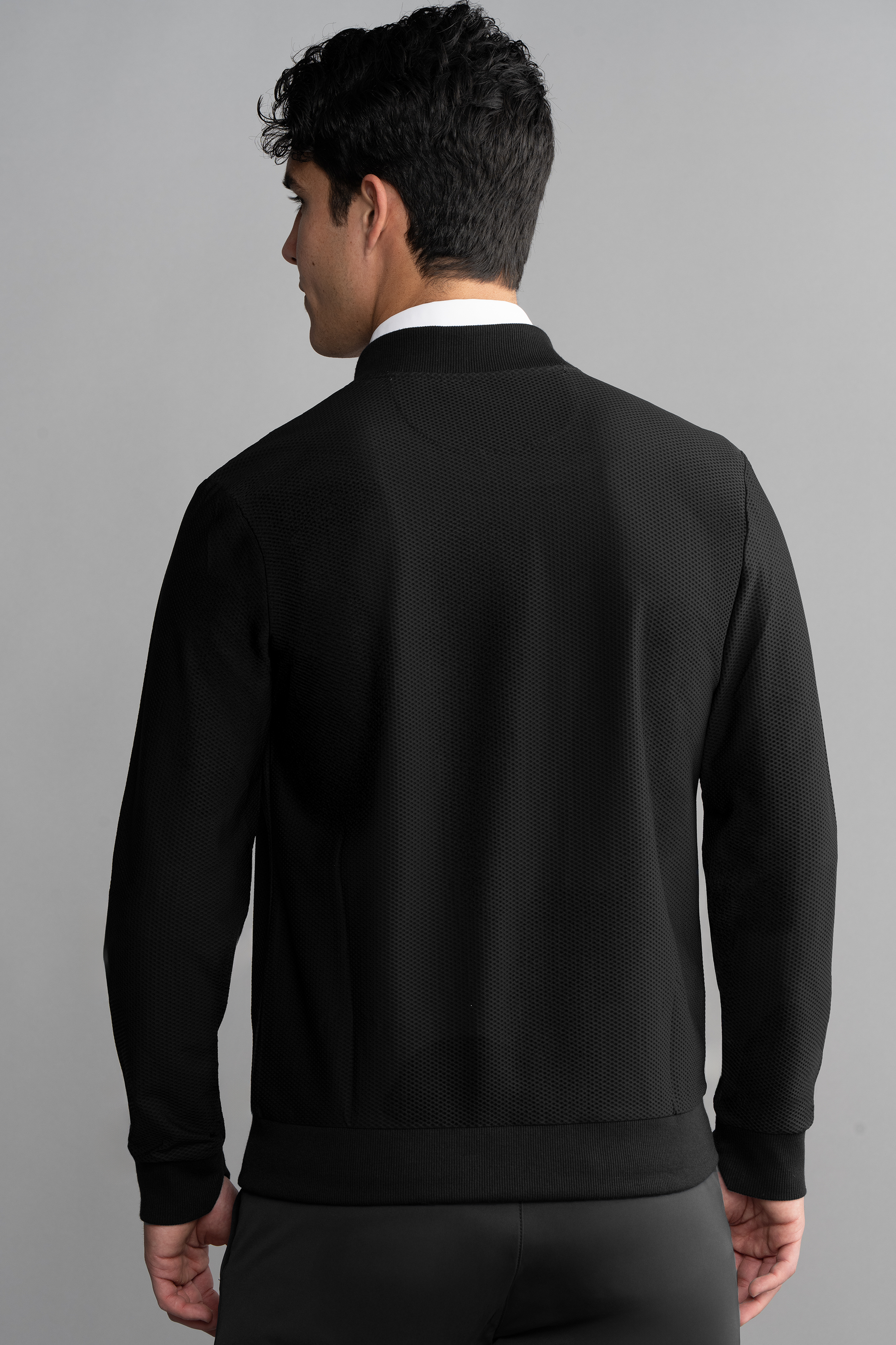 A man with short, dark curly hair wears the Twillory AIR Bomber in Black over a white shirt. This breathable jacket stands out against a plain light gray background as he turns slightly to the left.