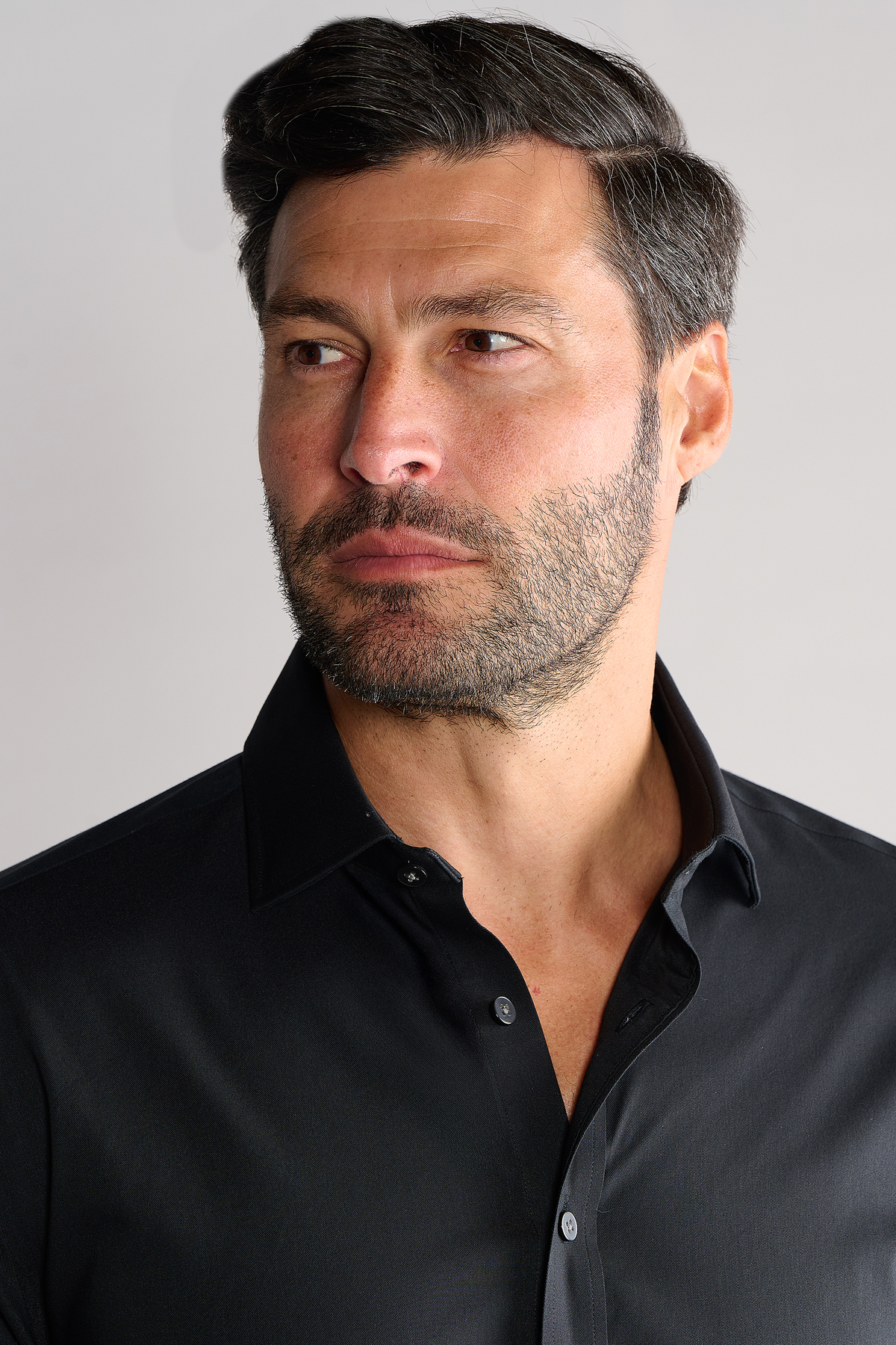 A man with dark hair and a trimmed beard wears the Twillory Leader Solid // Black stretch dress shirt. His collar is open, hair neatly styled, and he looks to the side with a neutral expression against a plain light background.