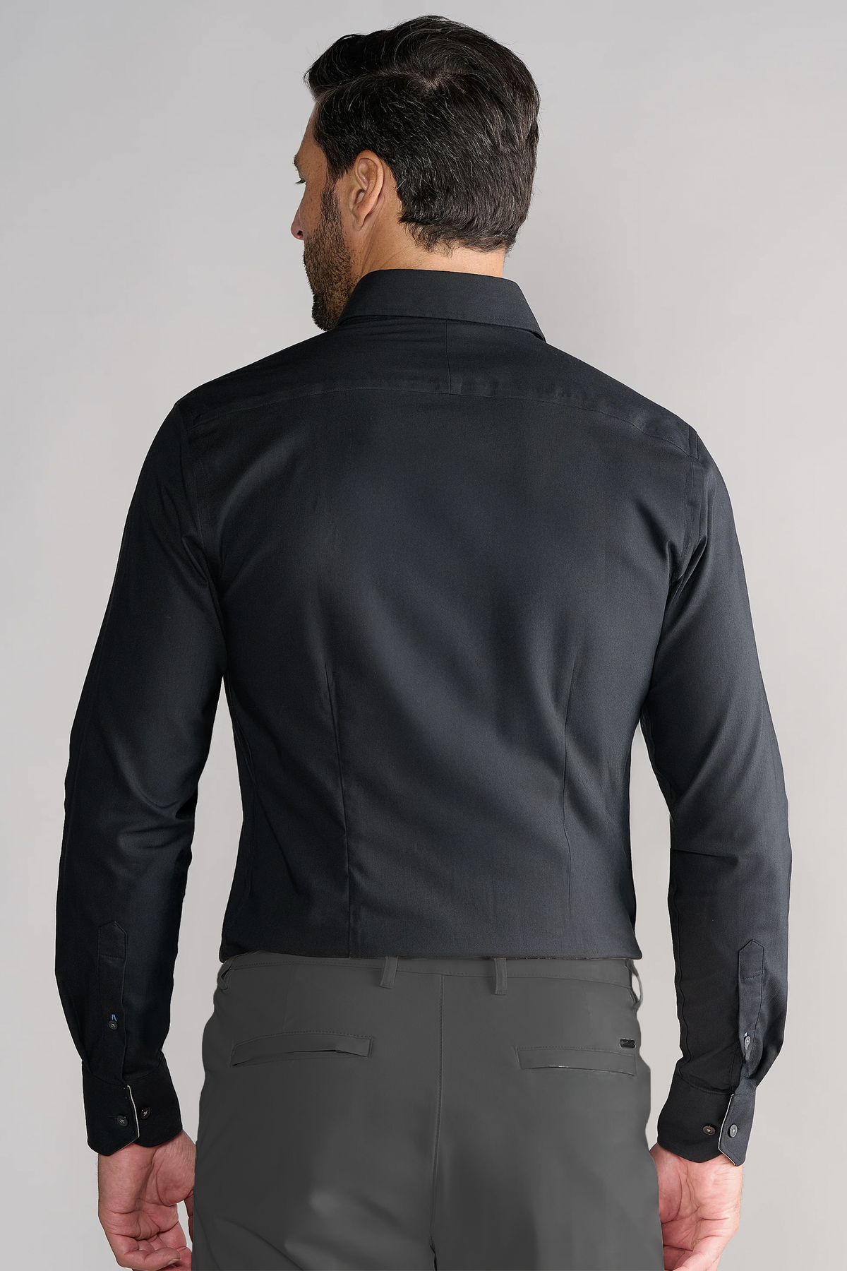A man with dark hair is seen from behind wearing the Twillory Leader Solid // Black stretch dress shirt and gray dress pants, arms at his sides, standing against a plain light gray background.