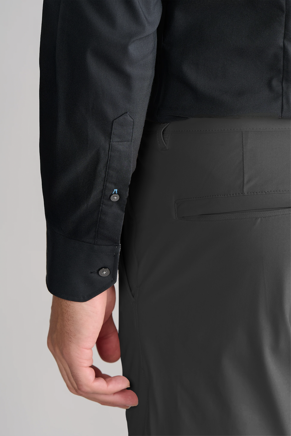 A close-up of a person’s left arm and hand, wearing Twillory’s Leader Solid // Black stretch dress shirt with buttoned cuffs and gray dress pants, highlights the clothing details against a light gray background.