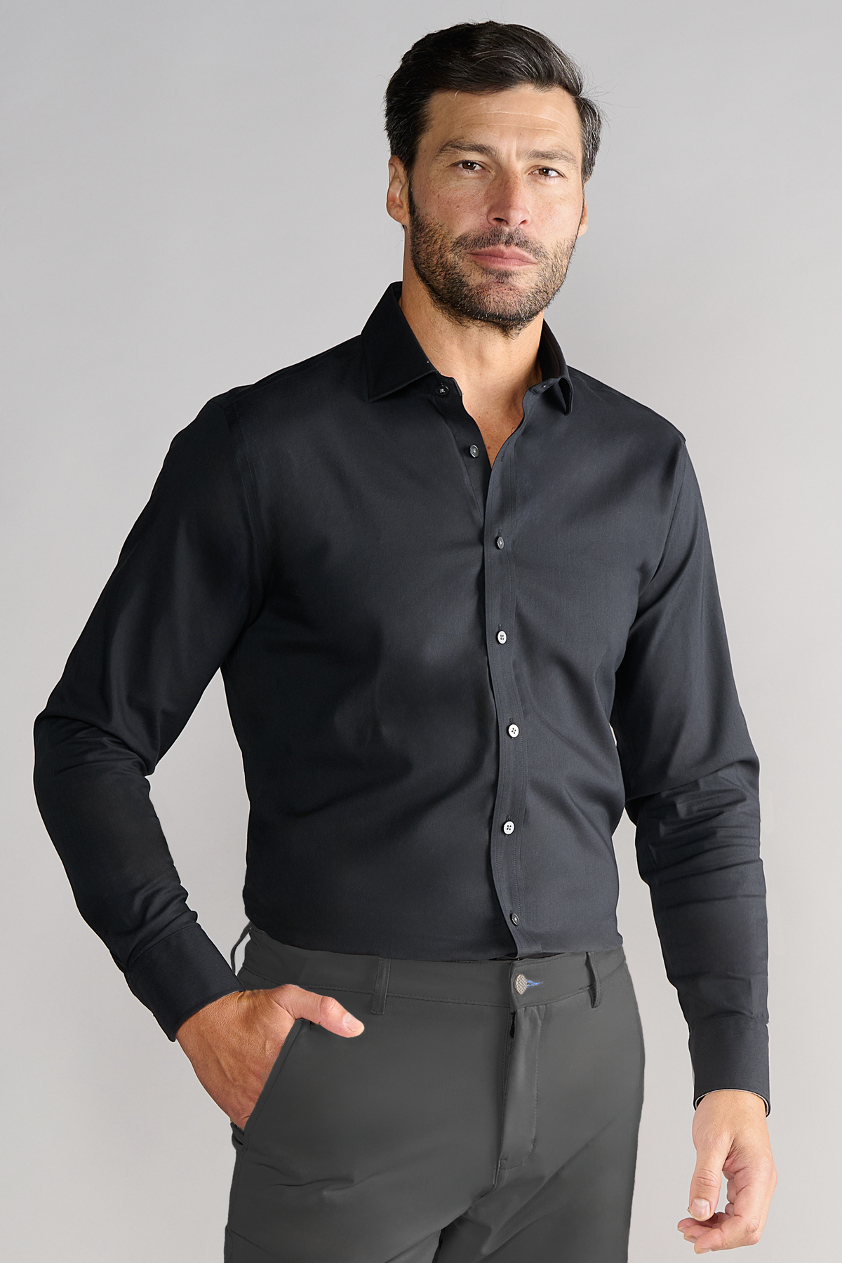A man with short dark hair and a trimmed beard stands against a plain light gray background, wearing Twillory's Leader Solid // Black fitted long-sleeve stretch dress shirt with buttoned cuffs and dark pants. One hand is in his pocket, expression neutral.