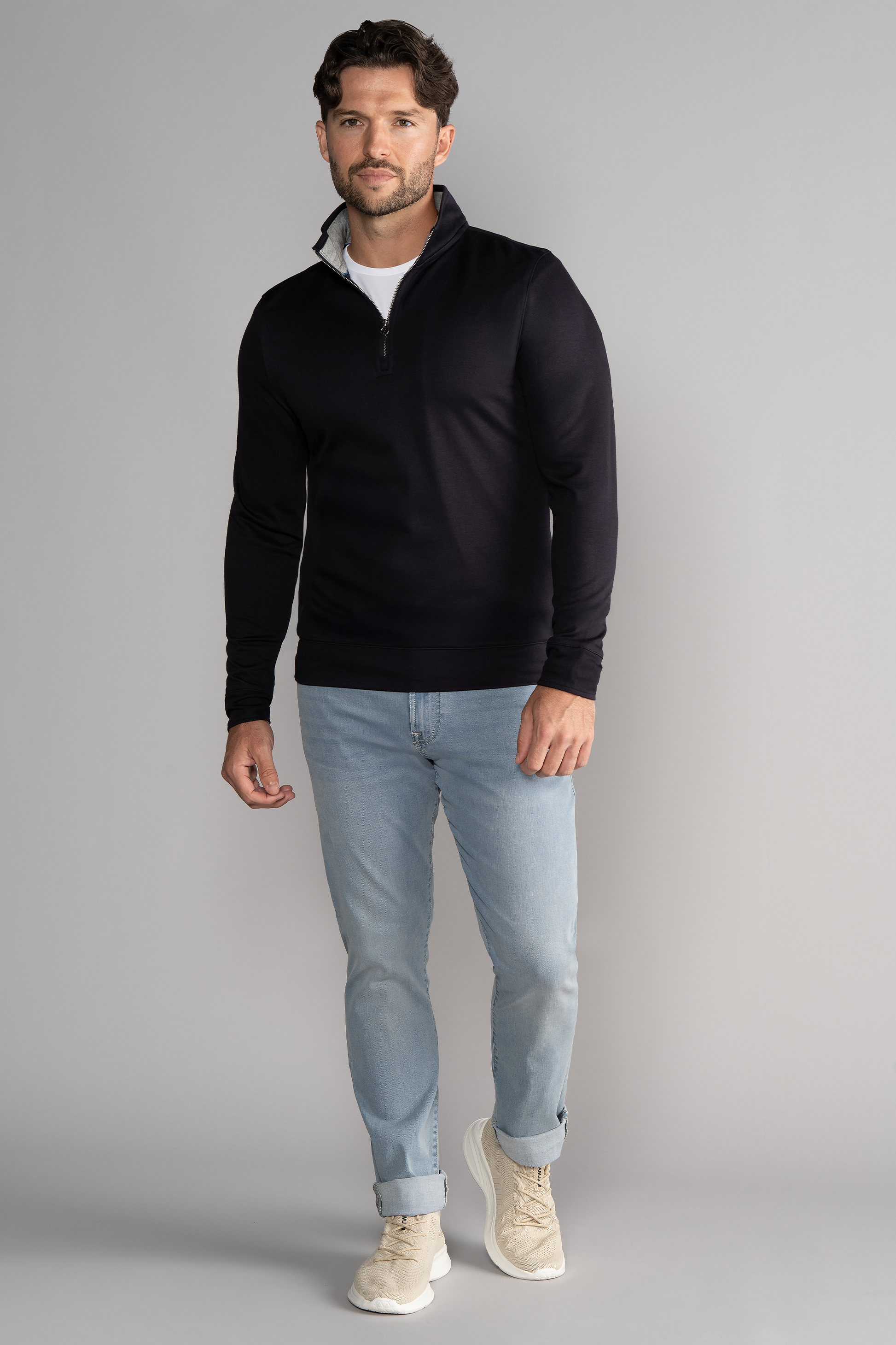 A man with short brown hair and a trimmed beard stands before a plain gray background, confidently wearing the Twillory Performance Quarter Zip in Black over a crisp shirt, light blue cuffed jeans, and beige sneakers while looking at the camera.