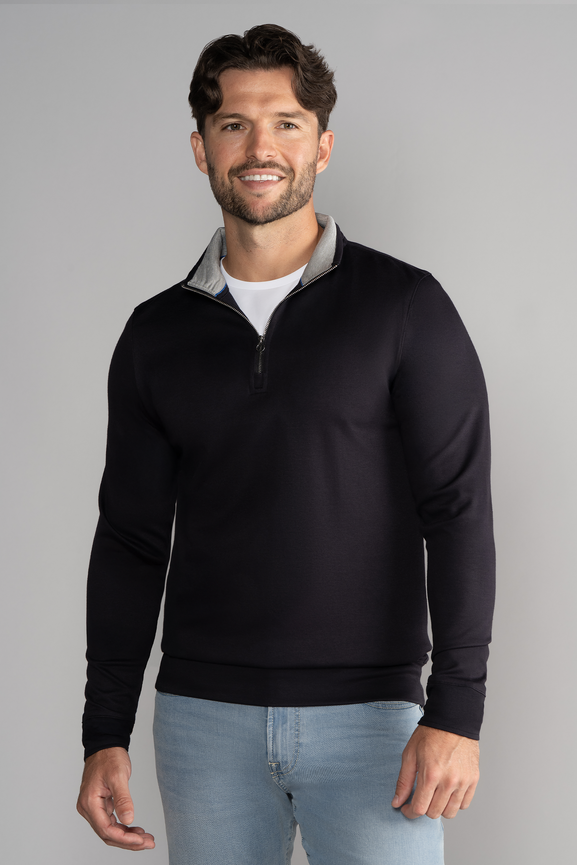 A man with short brown hair and a beard smiles against a gray background, wearing Twillory’s Performance Quarter Zip in black over a white Twillory shirt and light blue jeans. His right arm is slightly bent.