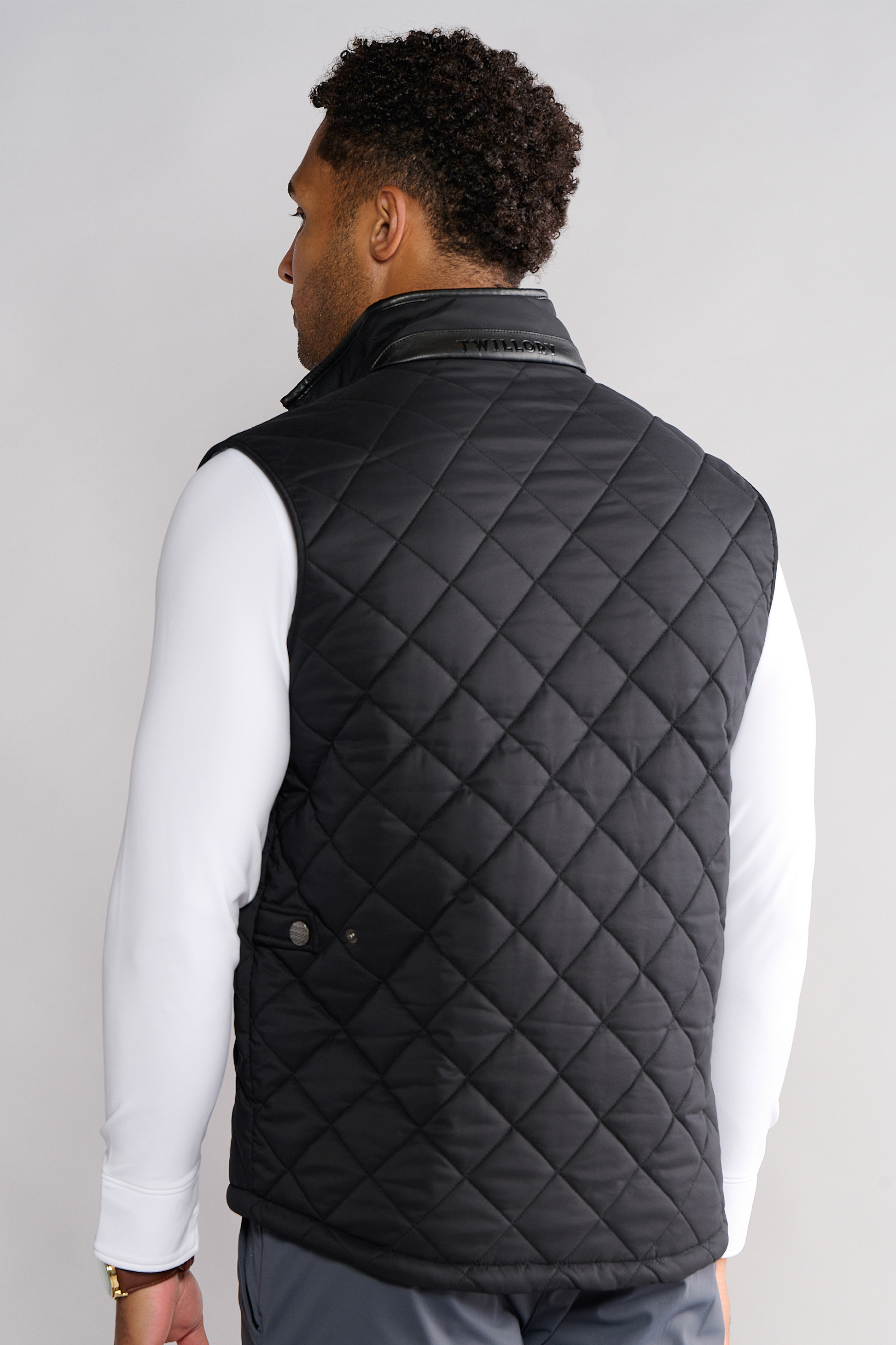A man faces away, wearing Twillory's Performance Quilted Vest—black, with a leather-trimmed collar, diamond pattern, snap waist buttons, and machine washability—over a white long-sleeve shirt. The background is plain light gray.