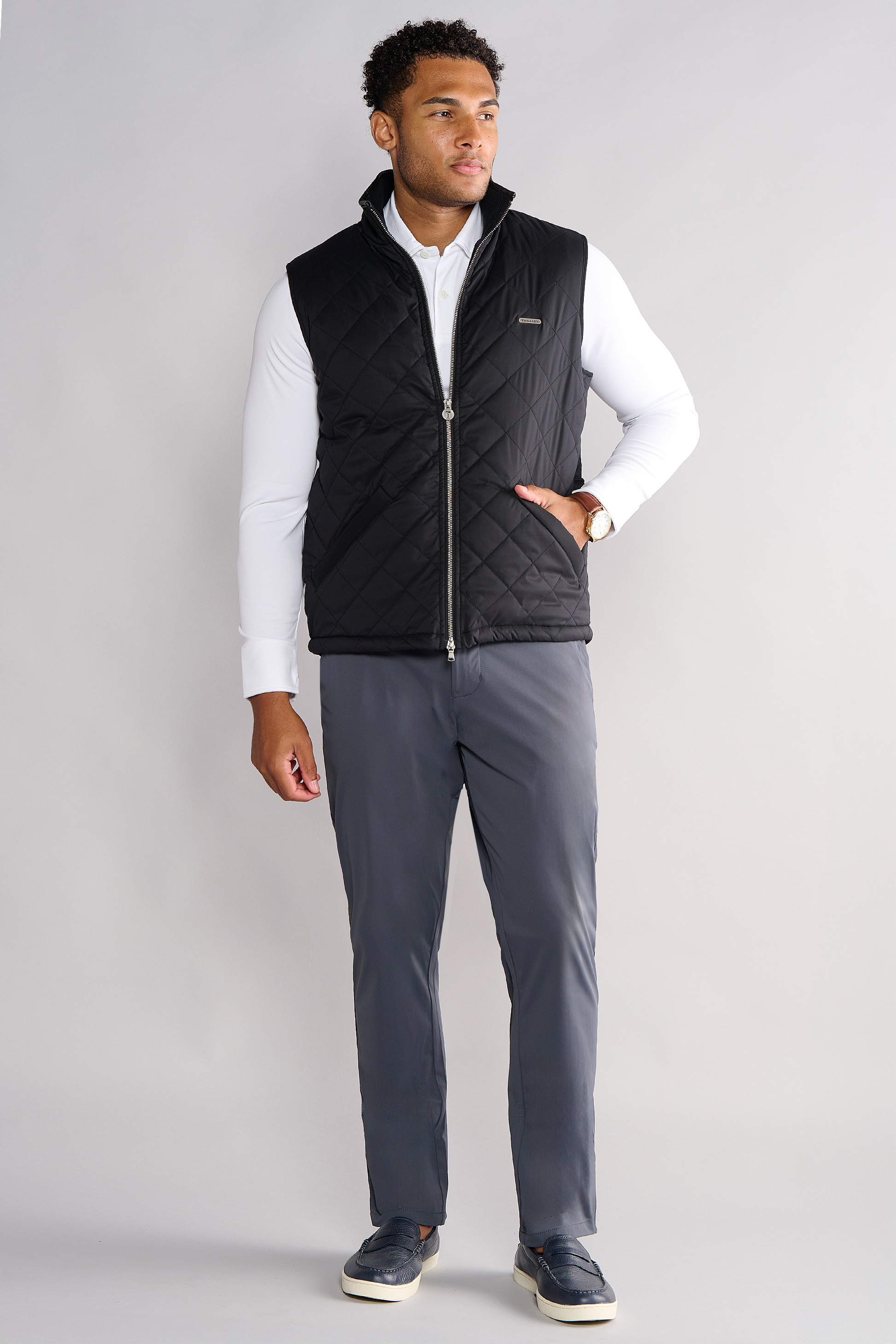 A man stands against a plain light gray background, wearing the Twillory Performance Quilted Vest over a white long-sleeve polo, gray pants, and black slip-on shoes. He looks slightly to the side with one hand in his pocket.