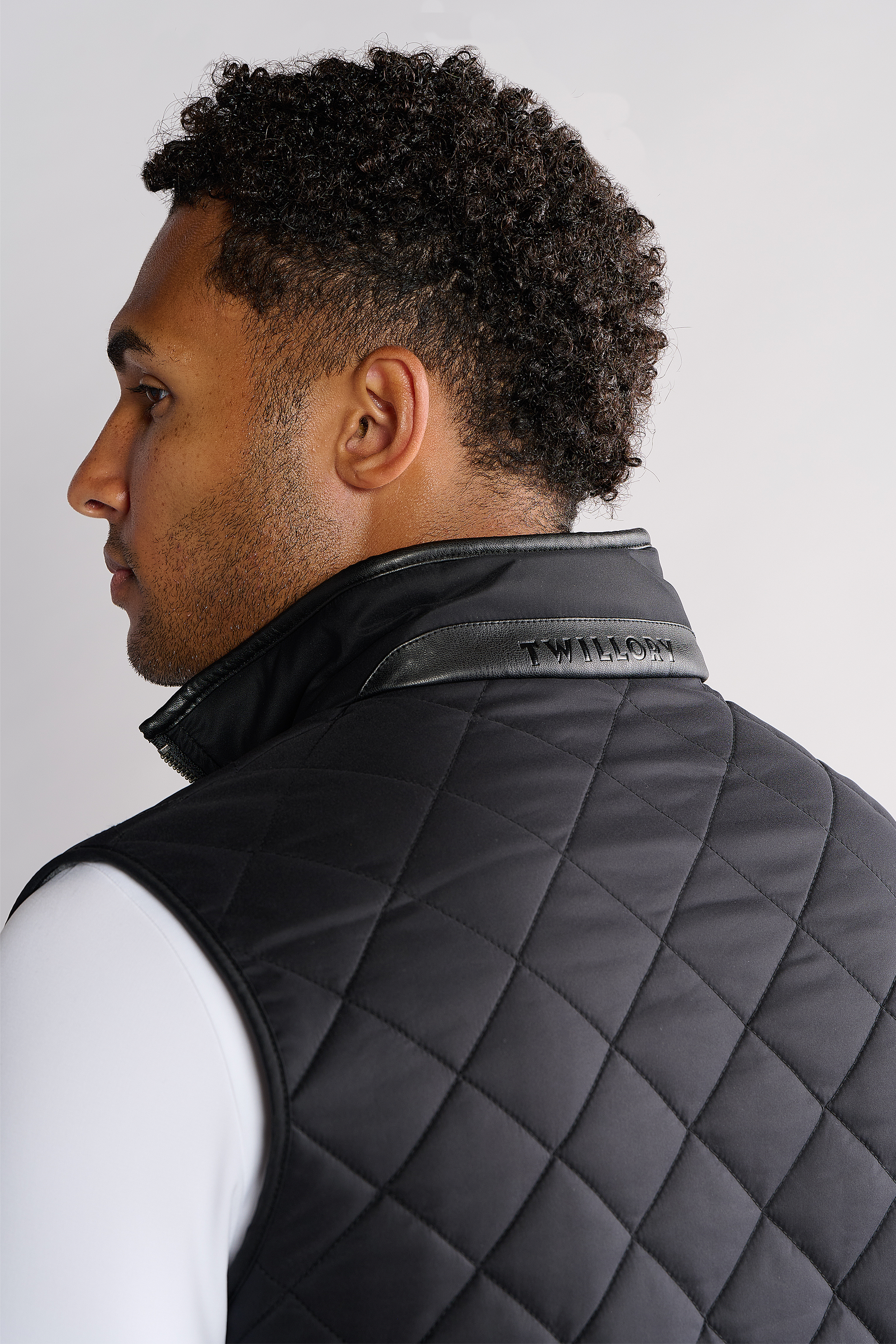 A man with short curly hair is seen from the back and side, wearing Twillory’s Performance Quilted Vest with a leather collar detail over a white long-sleeve shirt, set against a plain light gray background.