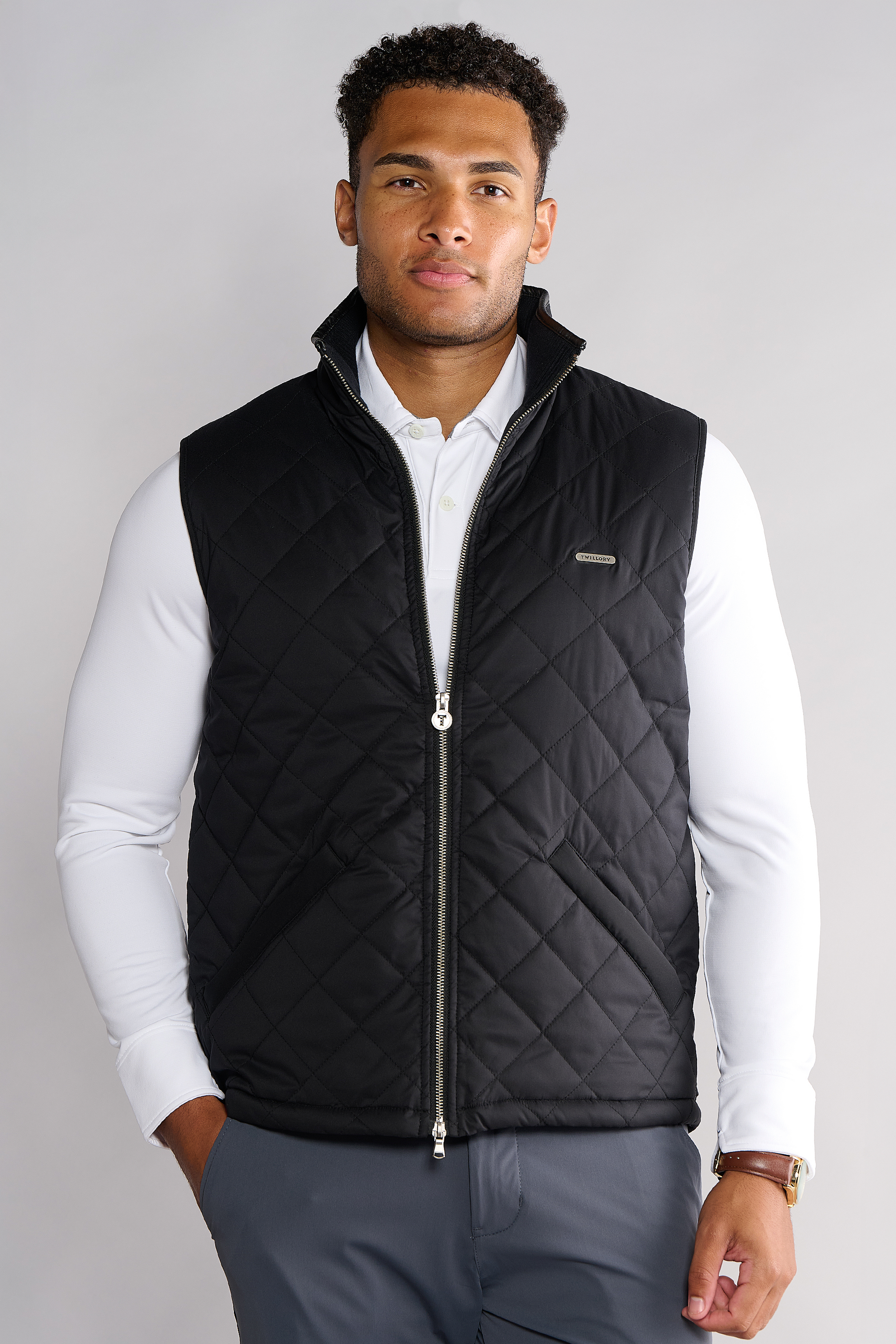 A man with short curly hair stands facing the camera, hands in pockets, wearing a Twillory Performance Quilted Vest with diamond stitching and chest logo over a white shirt and gray pants. The vest has Thinsulate fleece lining and is machine washable.
