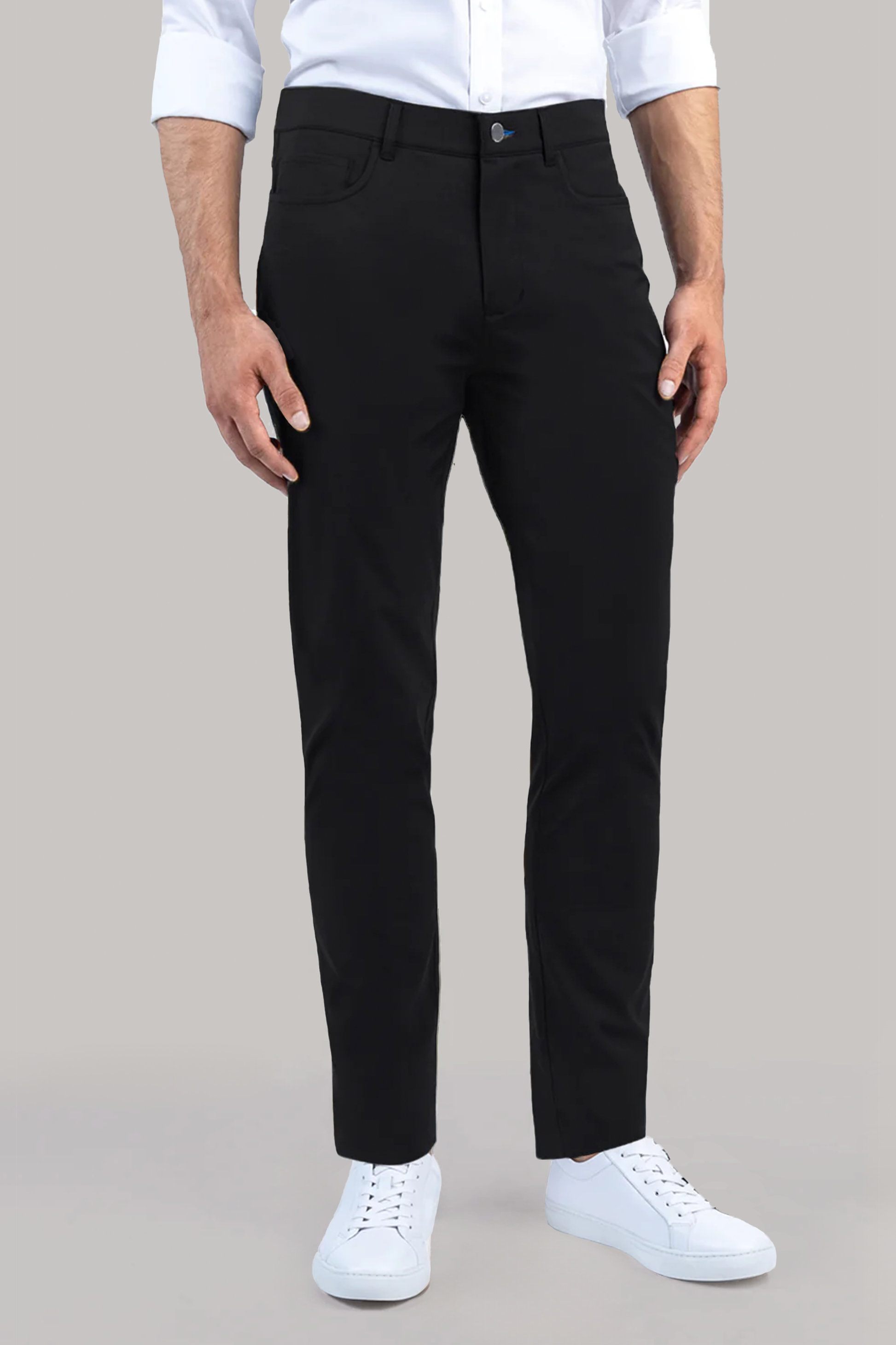 A man wears Performance Five Pocket // BLACK slim-fit pants and a white 4-way stretch shirt with rolled-up sleeves, paired with white sneakers, standing against a plain light grey background.