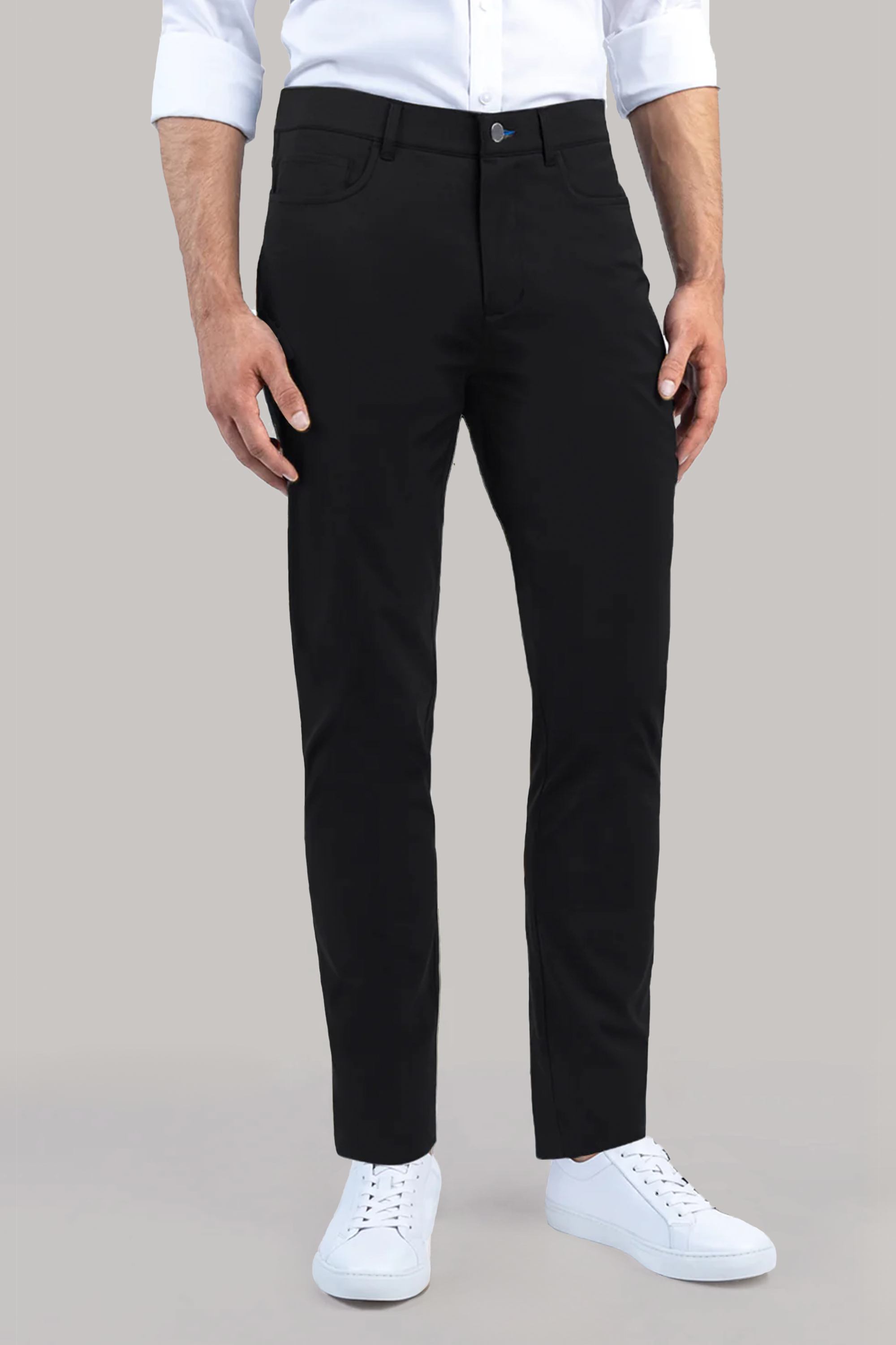 A man wears Performance Five Pocket // BLACK slim-fit pants and a white 4-way stretch shirt with rolled-up sleeves, paired with white sneakers, standing against a plain light grey background.