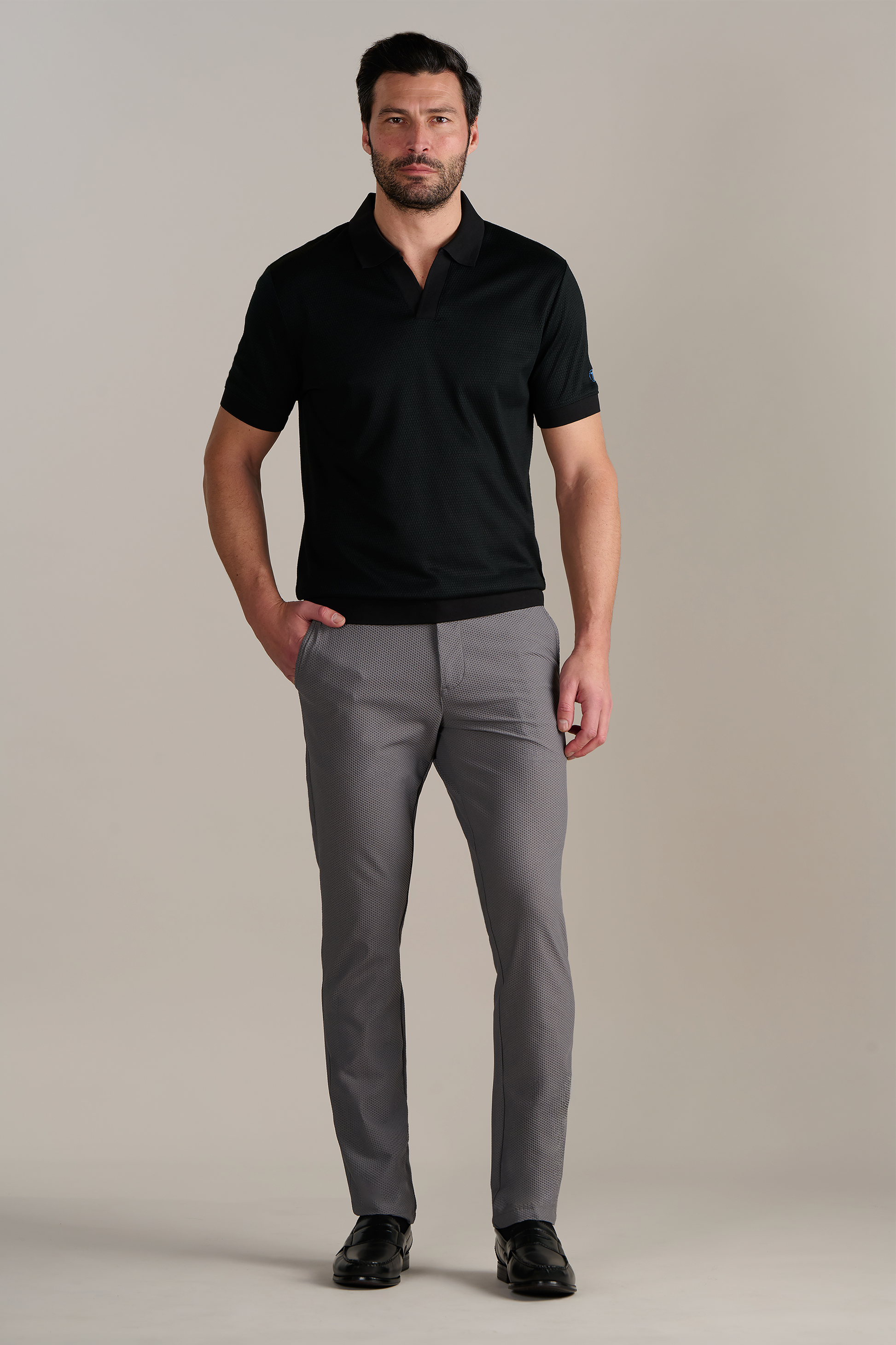 A man with short dark hair and a beard stands facing forward against a plain background, wearing Twillory's Short Sleeve Sweater Polo Solid in Black, light gray pants, and black dress shoes. His hands are in his pockets and he has a neutral expression.