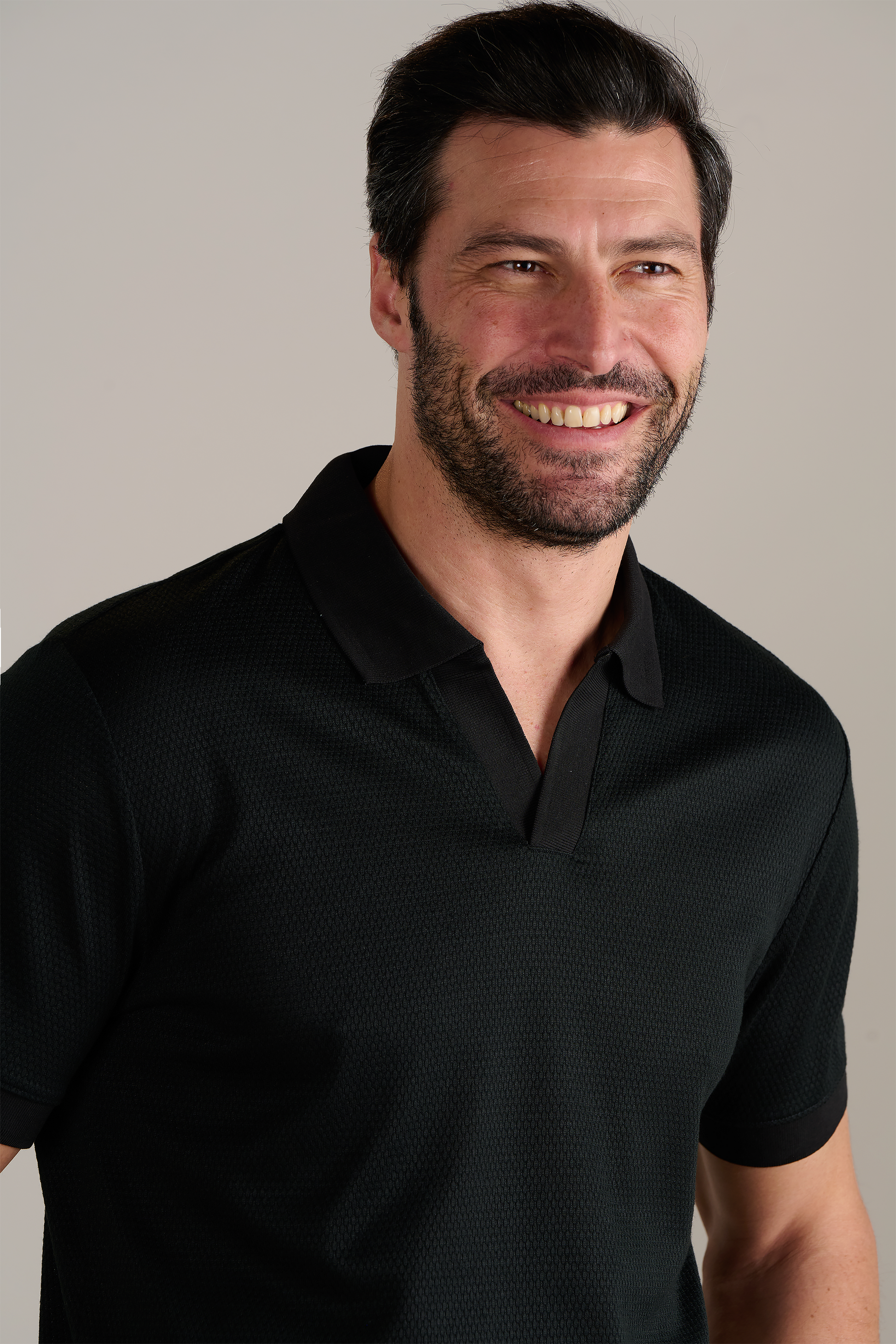 A smiling man with short dark hair and a beard wears the Twillory Short Sleeve Sweater Polo Solid in black, made from wrinkle-free performance fabric. He stands against a plain light gray background, facing slightly to the left.