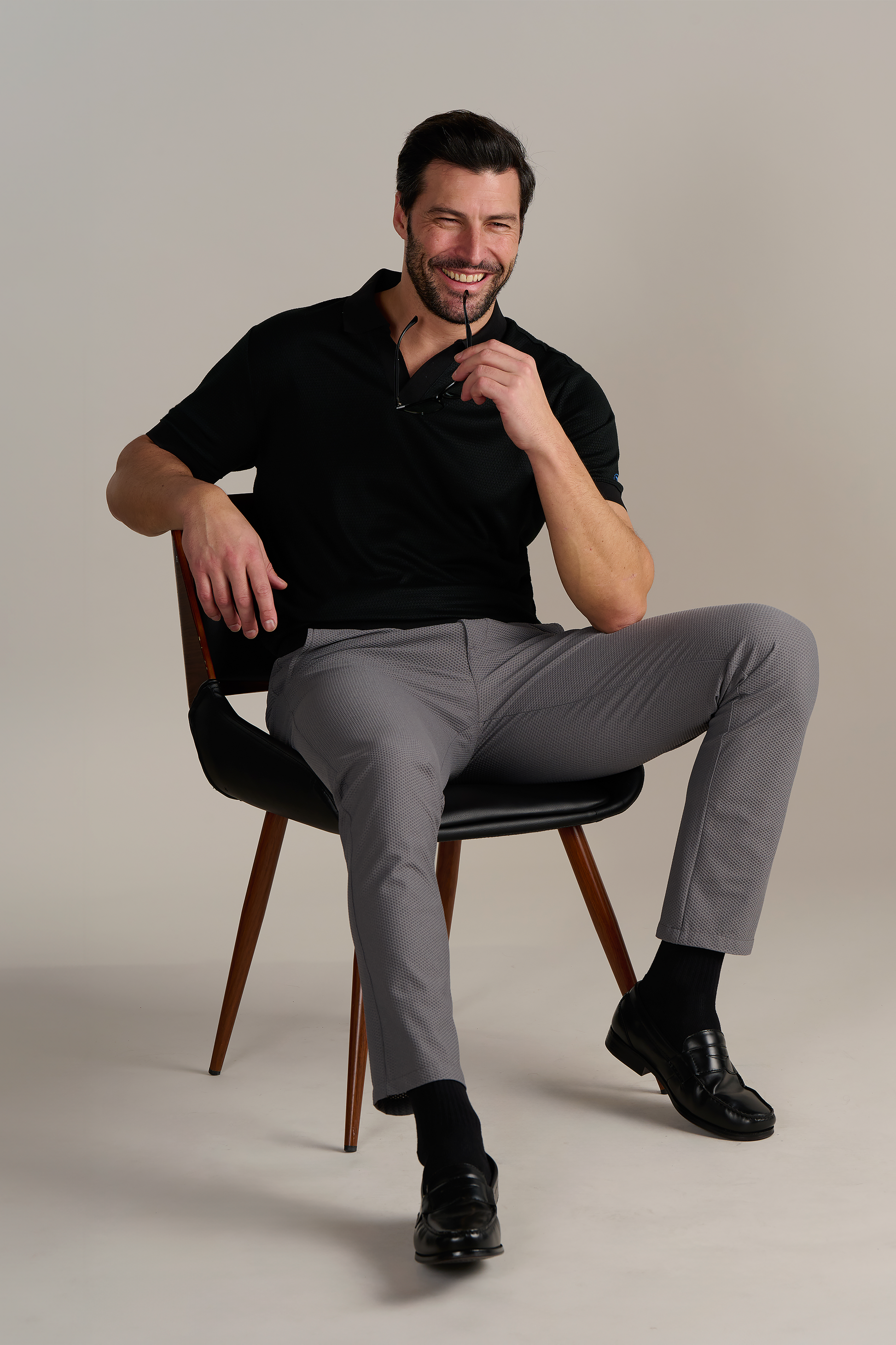 A smiling man with dark hair and a beard sits on a wooden chair, wearing Twillory's Short Sleeve Sweater Polo Solid in black, gray pants, and black loafers. The plain light gray background highlights his wrinkle-free style.