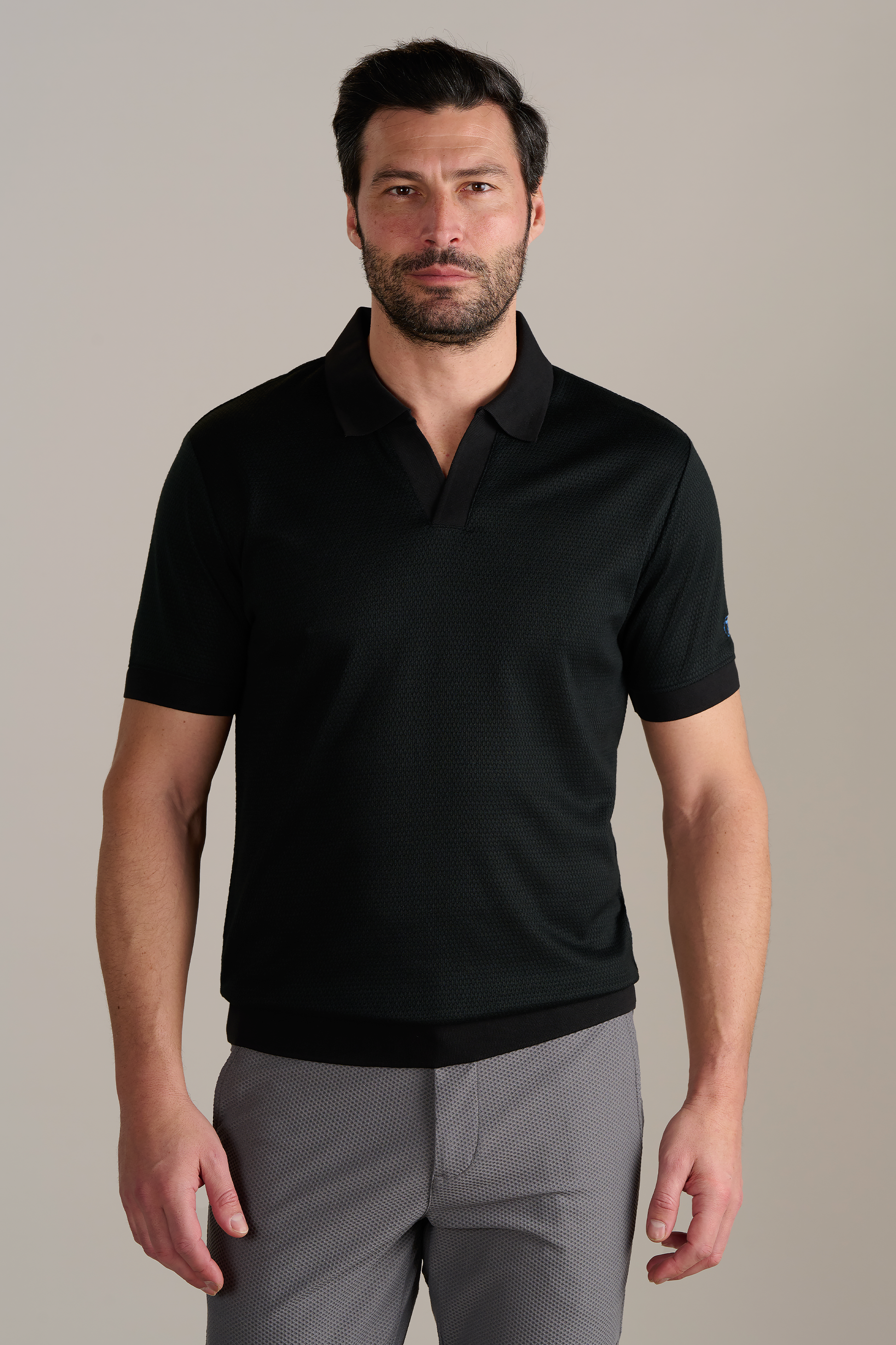 A man with short dark hair, a beard, and light skin stands facing forward in a relaxed pose. He wears the Twillory Short Sleeve Sweater Polo Solid in black and light gray wrinkle-free performance pants, set against a plain background.
