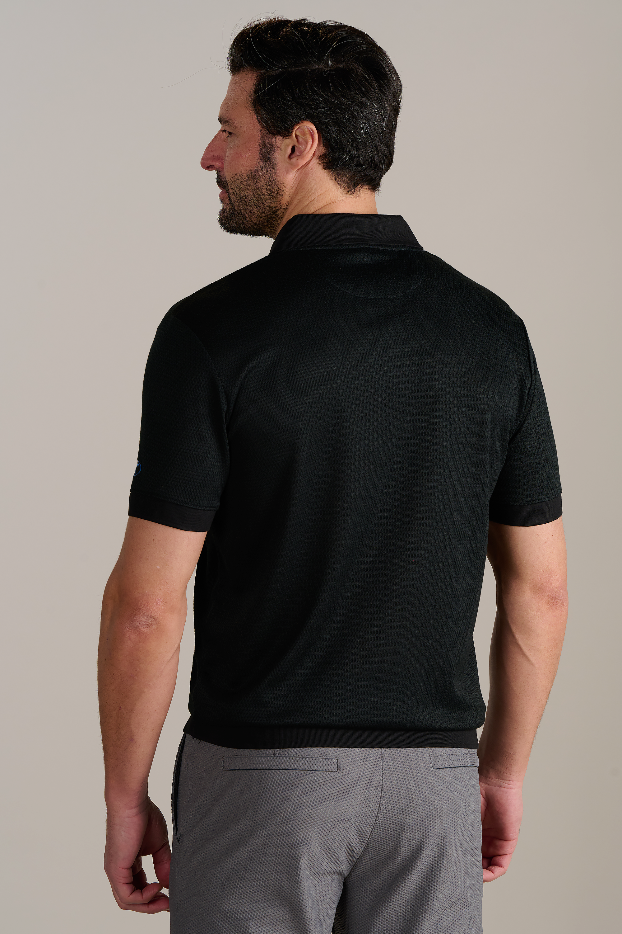 A man with short dark hair and a beard faces away, wearing Twillory’s Short Sleeve Sweater Polo Solid in black and gray pants. He stands against a plain light background, left arm relaxed and right arm slightly bent.
