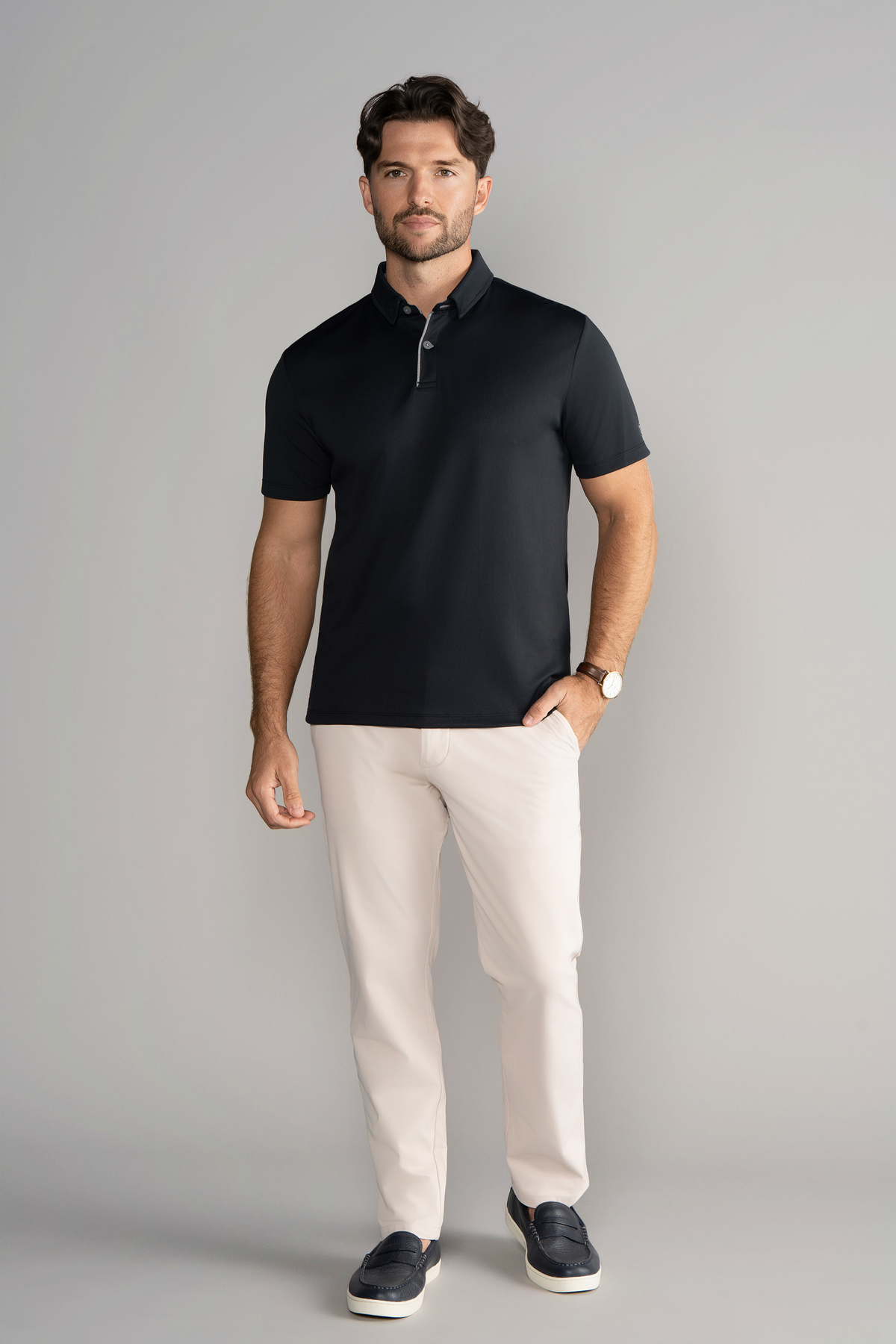 A man with short brown hair and a beard stands facing forward against a light gray background, wearing the Twillory Short Sleeve Polo Contrast in Black Grey, white pants, dark slip-on shoes, and a watch.