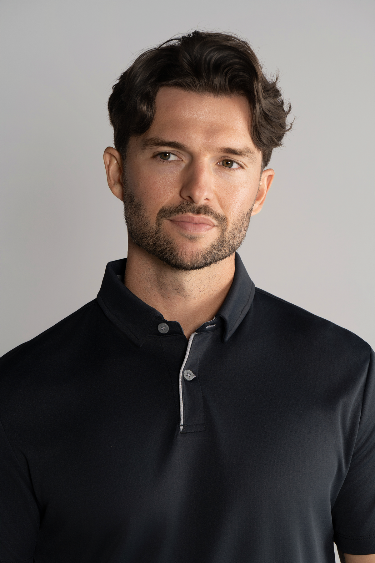 A man with wavy brown hair and a trimmed beard wears the Twillory Short Sleeve Polo Contrast in Black Grey. He faces the camera with a subtle smile against a plain light gray background.