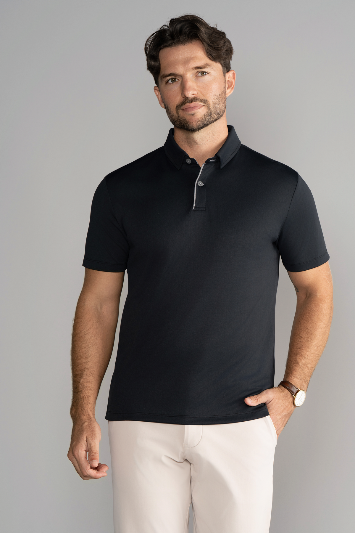 A man with short brown hair and a trimmed beard wears the Twillory Short Sleeve Polo Contrast in Black Grey and light beige pants, standing against a plain light gray background with a slight smile, wearing a wristwatch on his left wrist.