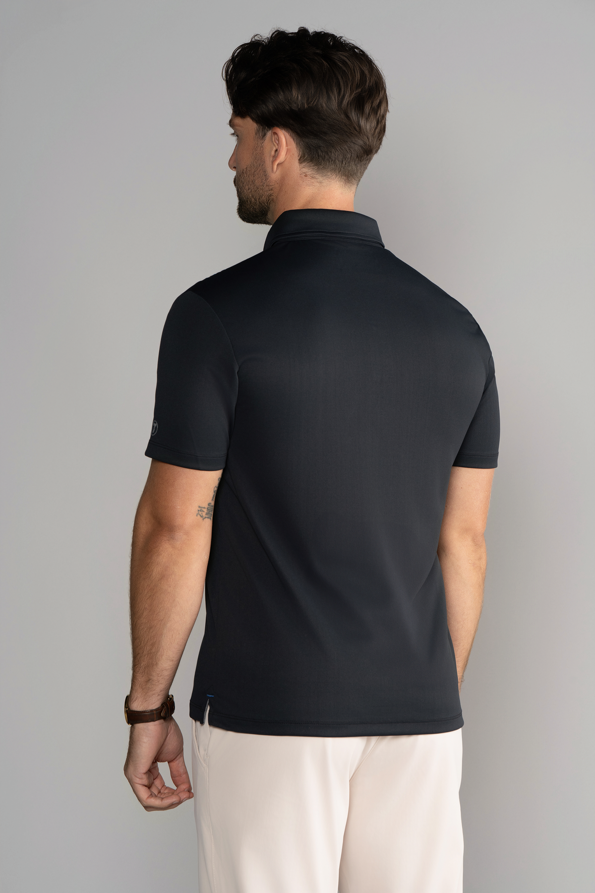 A man with short brown hair, seen from behind, wears the Twillory Short Sleeve Polo Contrast in Black Grey, featuring a tailored fit. He pairs it with light pants and accessories, while standing against a plain, softly lit background.