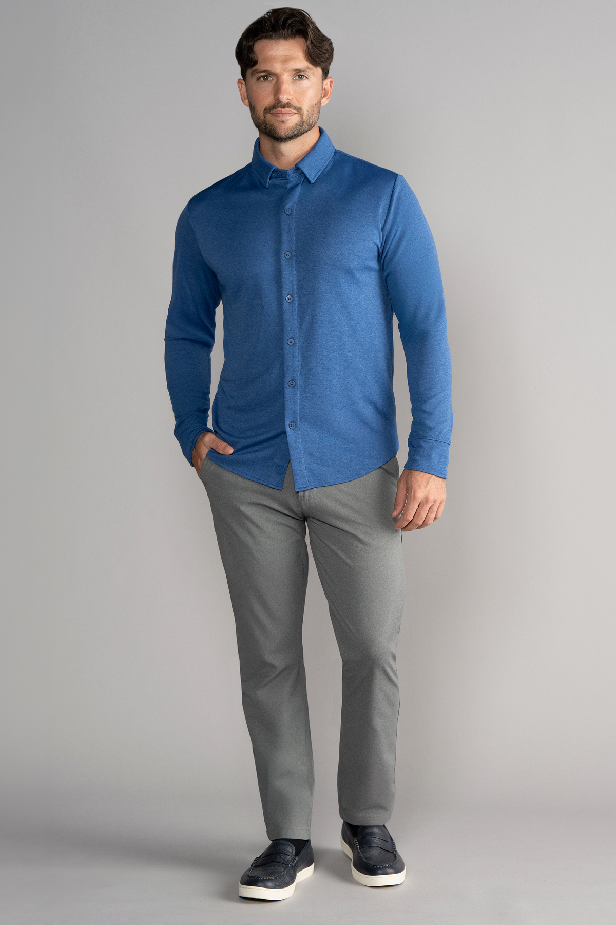 A man with short brown hair and a trimmed beard stands against a gray background, wearing the Twillory Long Sleeve Button Down Polo Solid in blue, light gray pants, and dark slip-on shoes. He poses confidently with one hand in his pocket.