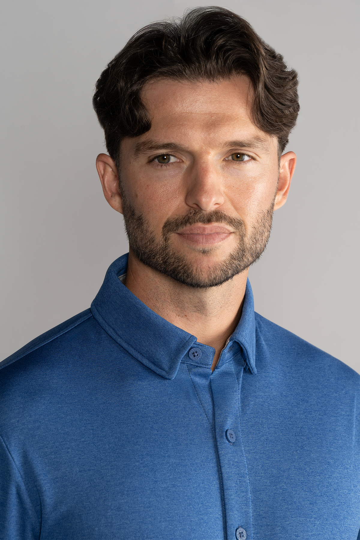 A man with wavy brown hair, light skin, and a trimmed beard faces the camera, wearing the Twillory Long Sleeve Button Down Polo Solid in blue. He has a neutral expression against a plain light gray background. Only his upper torso and head are visible.