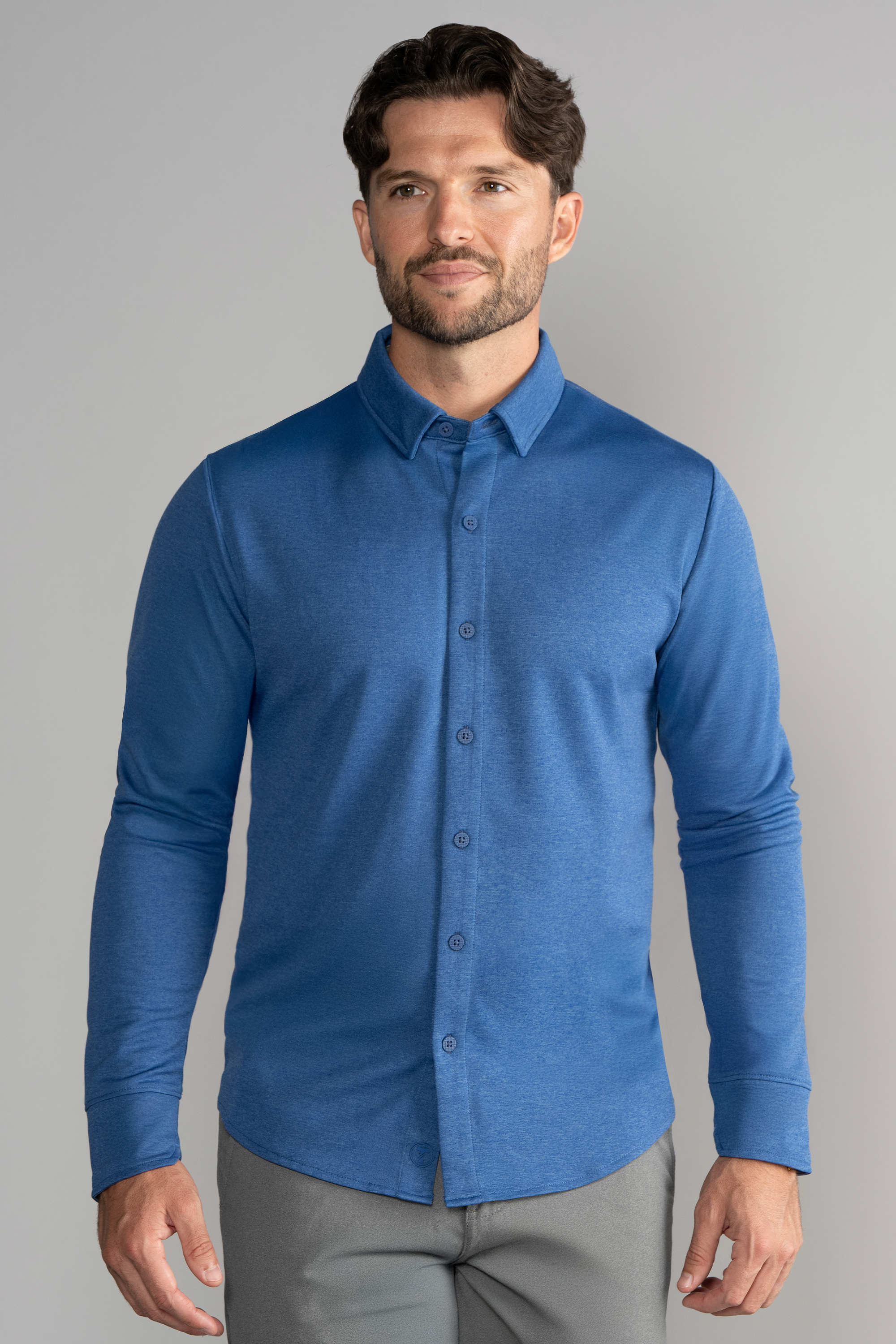 A man with short brown hair and a trimmed beard wears the Twillory Long Sleeve Button Down Polo Solid in blue and light gray pants, standing facing forward with arms relaxed against a plain light gray background.