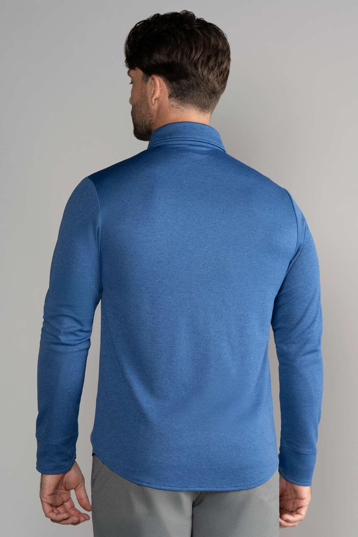 A man with short brown hair is seen from the back wearing Twillory's Long Sleeve Button Down Polo Solid in blue, paired with gray pants, standing against a plain light gray background.