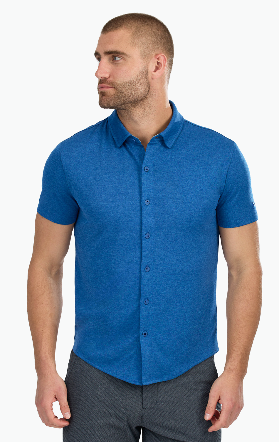 A man with short hair and a beard wears the Twillory Short Sleeve Button Down Polo Solid in blue and dark pants. He stands slightly turned left, looking away from the camera, arms relaxed against a plain white background.