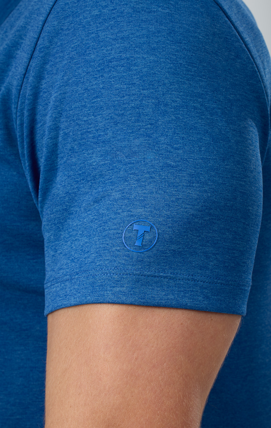 A close-up of a person’s upper arm wearing Twillory’s Short Sleeve Button Down Polo Solid in blue showcases a small circular logo by the sleeve hem. This moisture-wicking polo keeps you cool and comfortable against a neutral background.