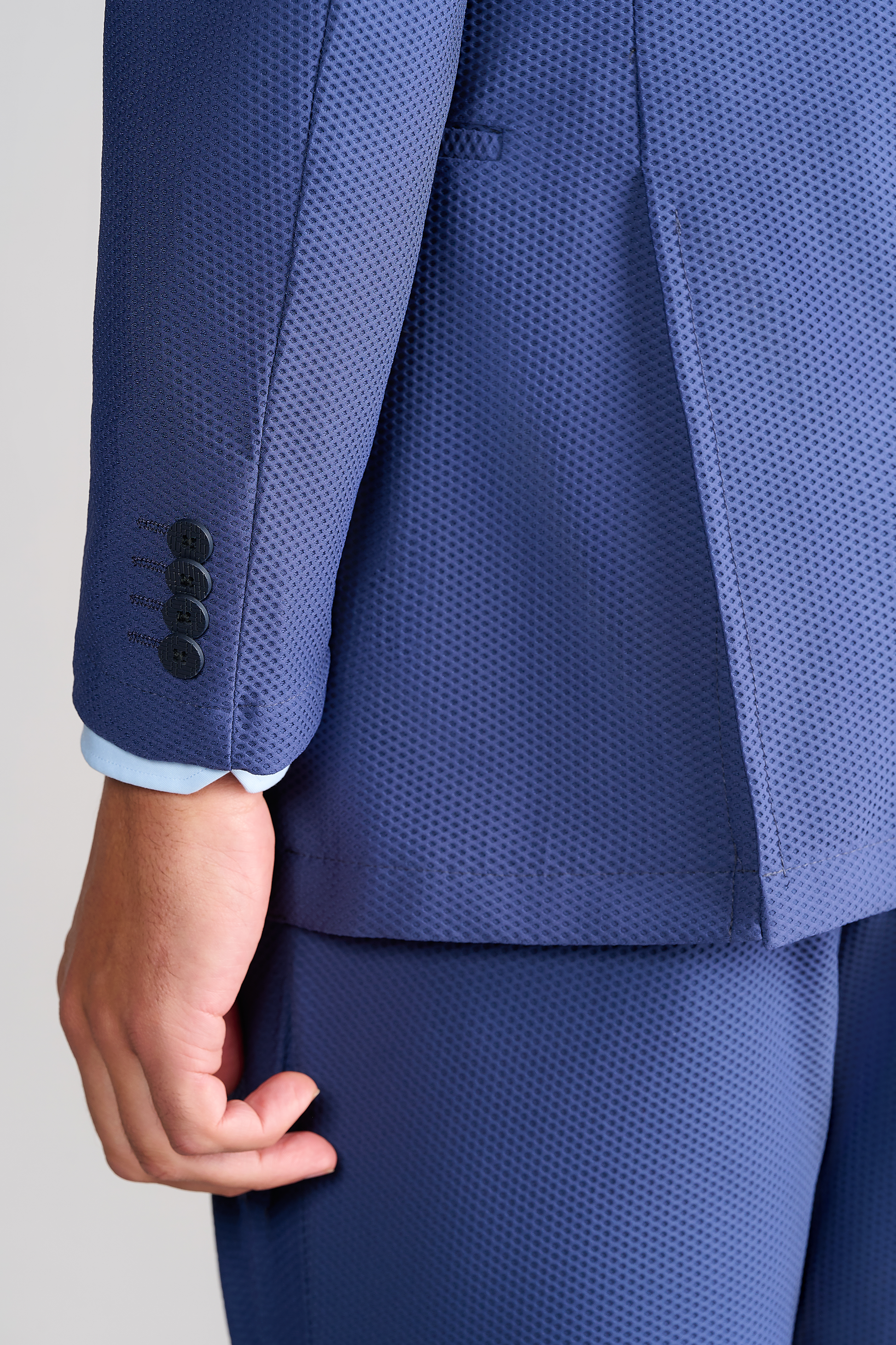 A close-up of a person’s left arm wearing the Twillory AIR Blazer // Blue. The textured fabric features a small dotted pattern, black sleeve buttons, and reveals a light blue shirt cuff at the wrist as their hand rests relaxed by their side.