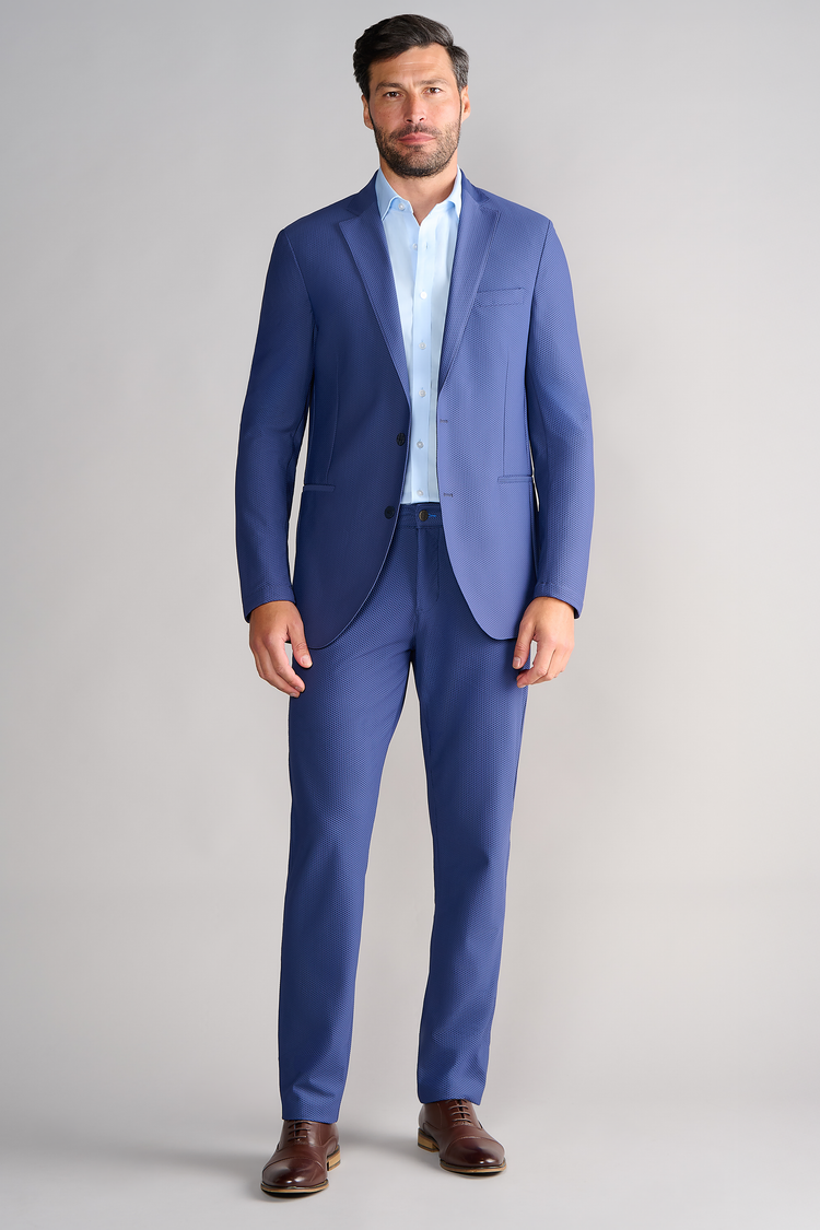 A man with short dark hair and a beard wears the Twillory AIR Blazer // Blue over a light blue dress shirt, no tie, and brown dress shoes. He stands facing forward against a plain light gray background with arms relaxed by his sides.