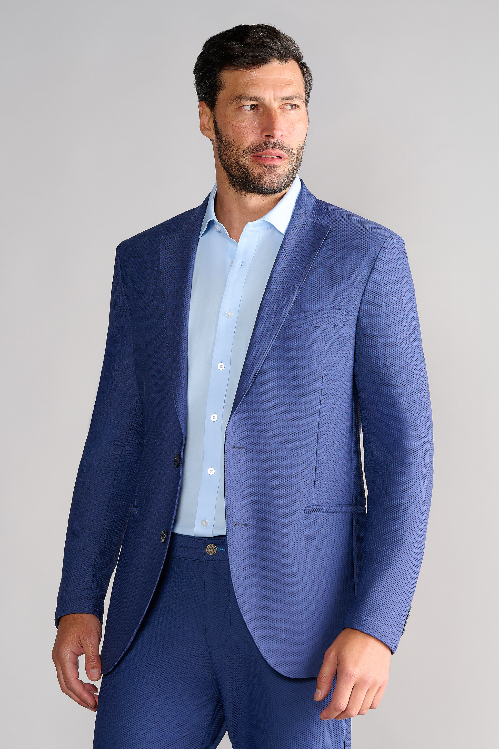 A man with short dark hair and a beard wears the Twillory AIR Blazer // Blue over a light blue dress shirt. The blazer’s 360° airflow fabric is textured, and he stands against a plain light gray background, gazing left neutrally.