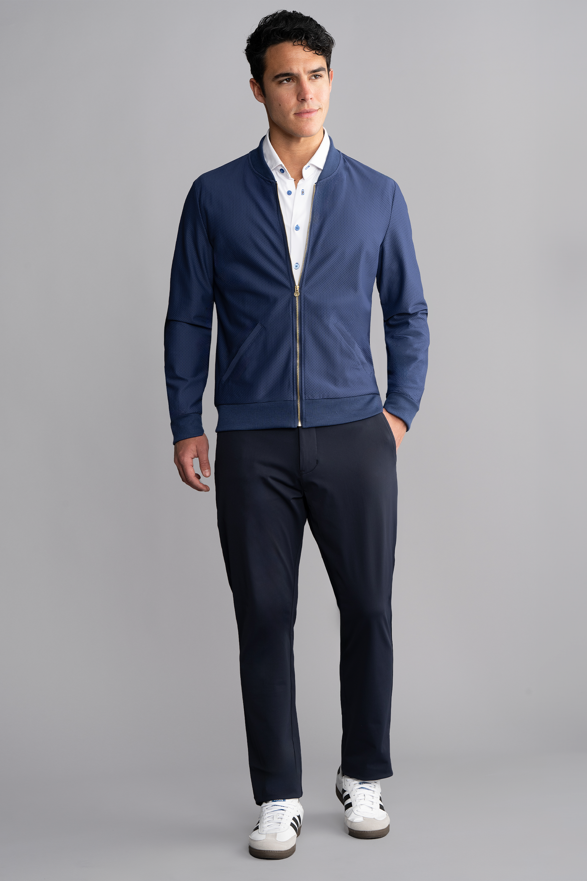 A man stands against a plain grey background wearing the Twillory AIR Bomber // Blue over a white button-down shirt, dark trousers, and white sneakers with black stripes. He looks slightly to the side with a neutral expression, left hand in his pocket.