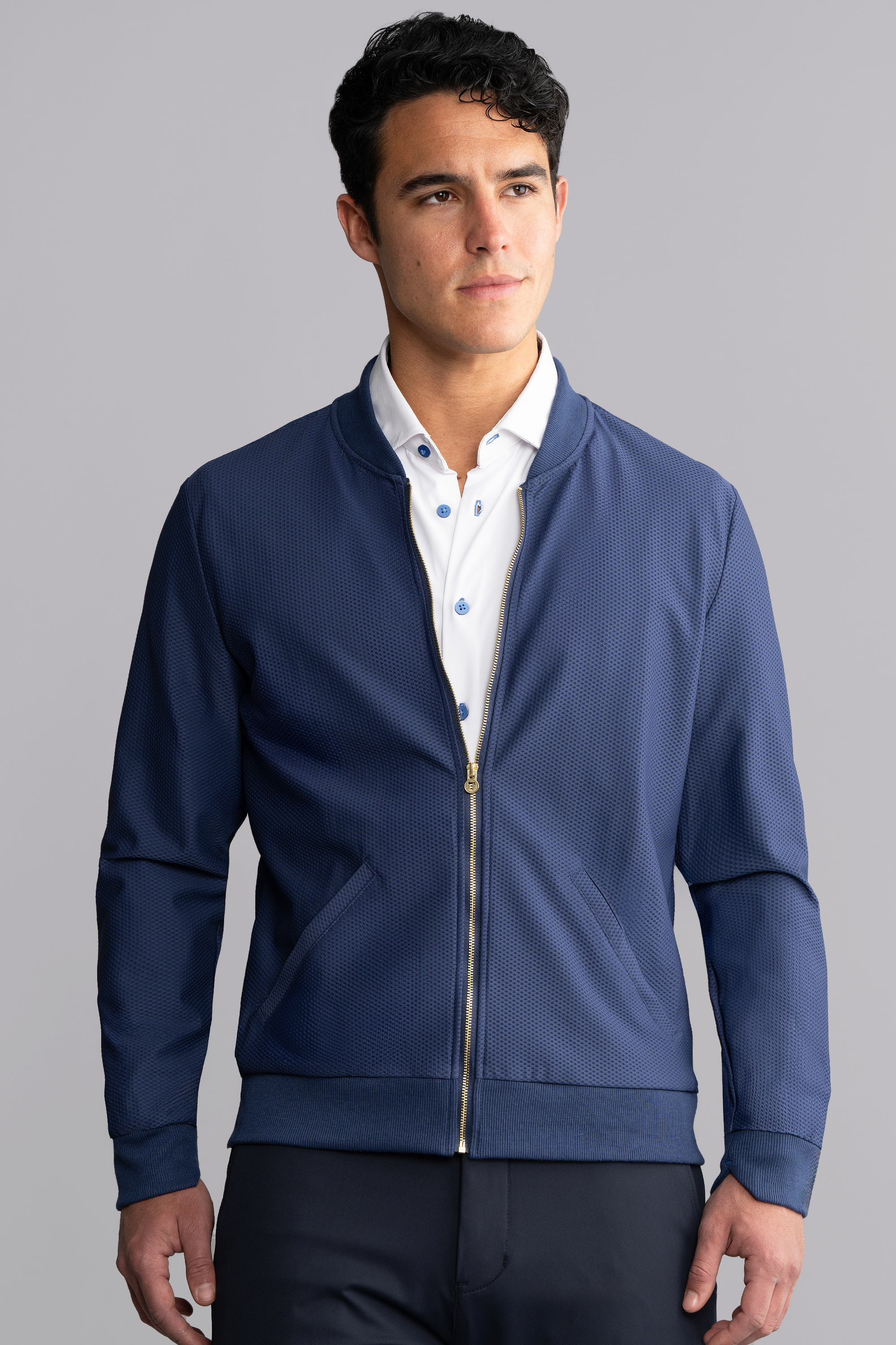 A man with short dark hair wears the Twillory AIR Bomber in blue over a white button-down shirt. He stands against a plain gray background, looking to the side with a neutral expression and his hands relaxed by his sides.
