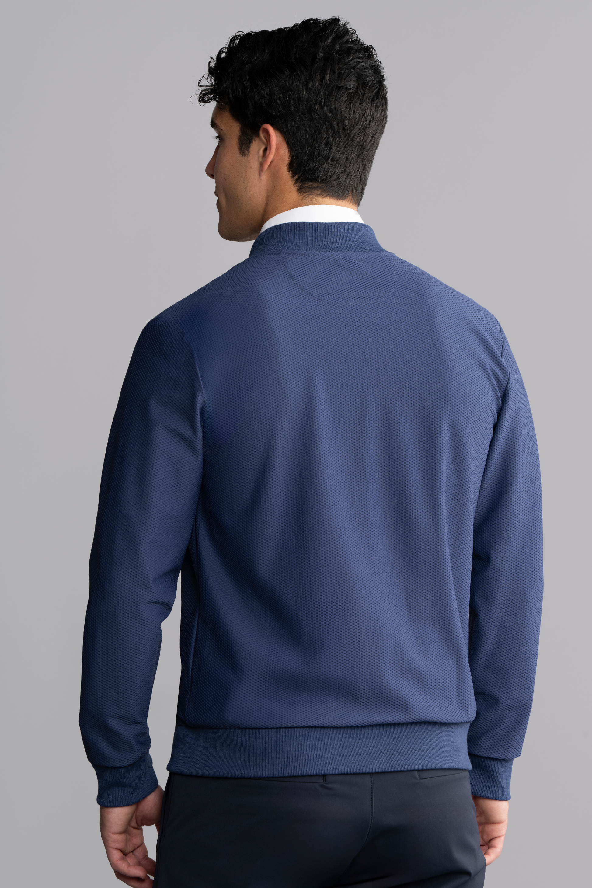 A man with short, curly black hair is seen from behind wearing the Twillory AIR Bomber // Blue, made with 360° Airflow Fabric for breathability. He stands against a plain light gray background, his head turned slightly left.