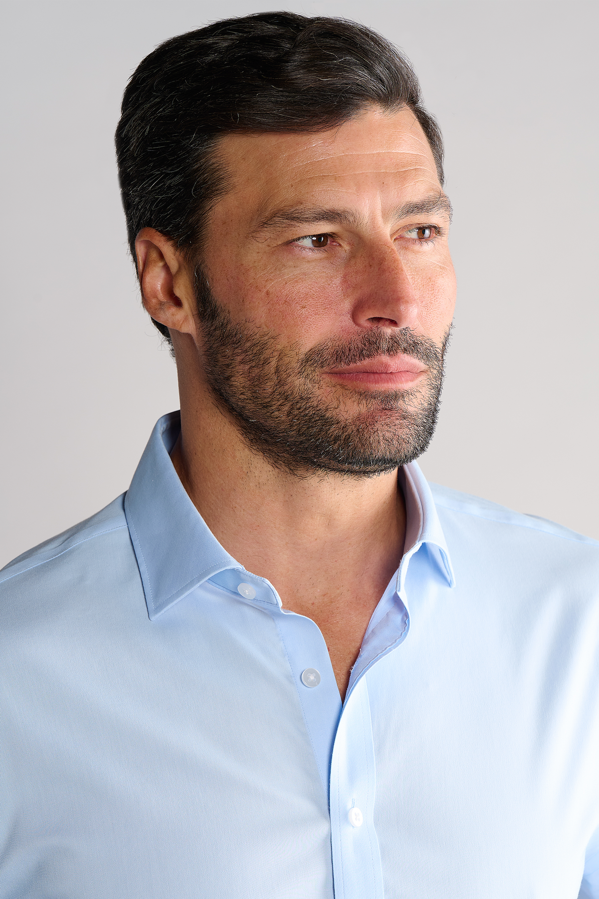 A man with short dark hair and a trimmed beard looks off to the side, wearing the Twillory Leader Solid // Blue non-iron shirt with the top button undone against a clean, light-colored background.