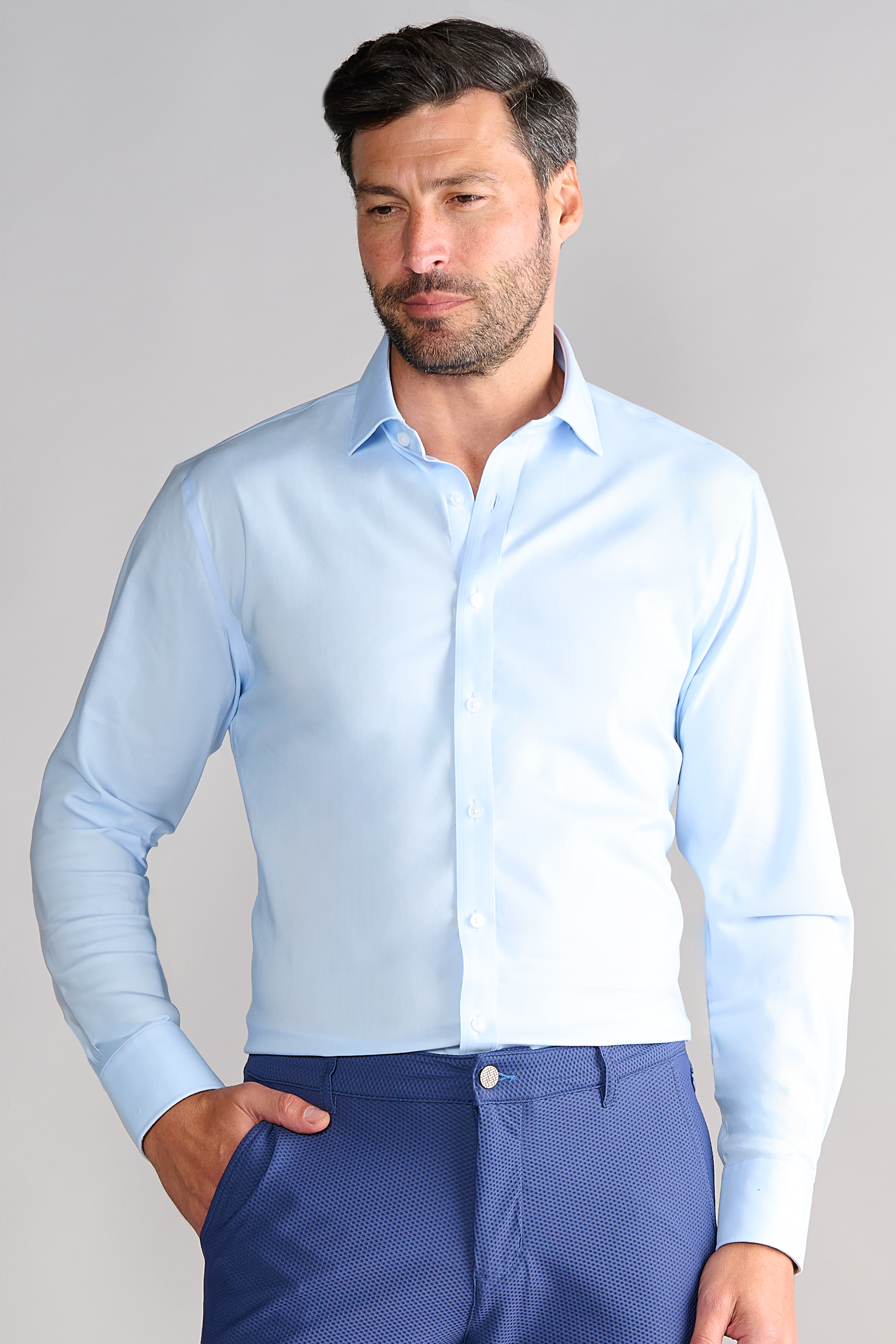 A man wears the Twillory Leader Solid // Blue non-iron shirt, tucked into patterned trousers. He stands against a plain gray background, looking slightly down and to the side with one hand in his pocket.