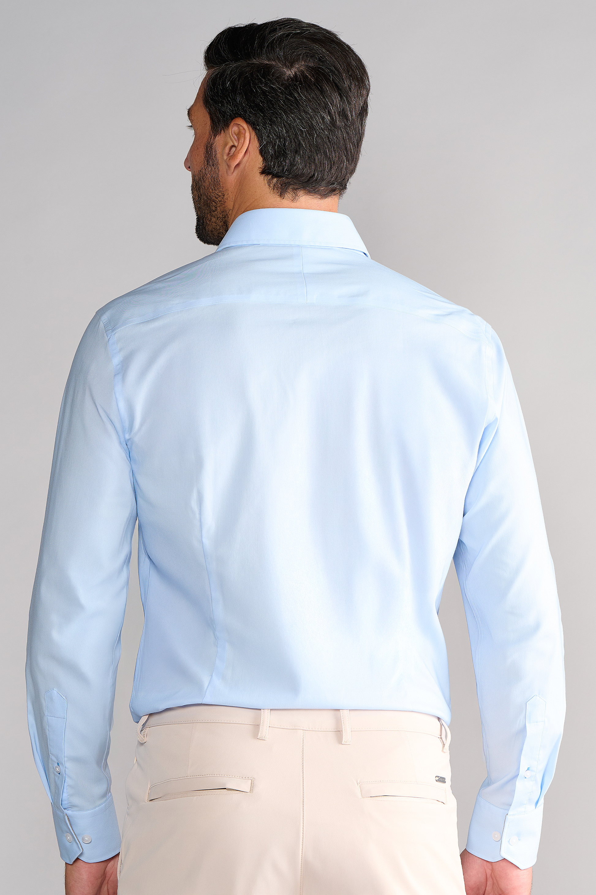 A man faces away in a tucked-in Twillory Leader Solid // Blue non-iron, long-sleeve dress shirt and beige pants. The plain light gray background contrasts with his short dark hair and neatly groomed beard.