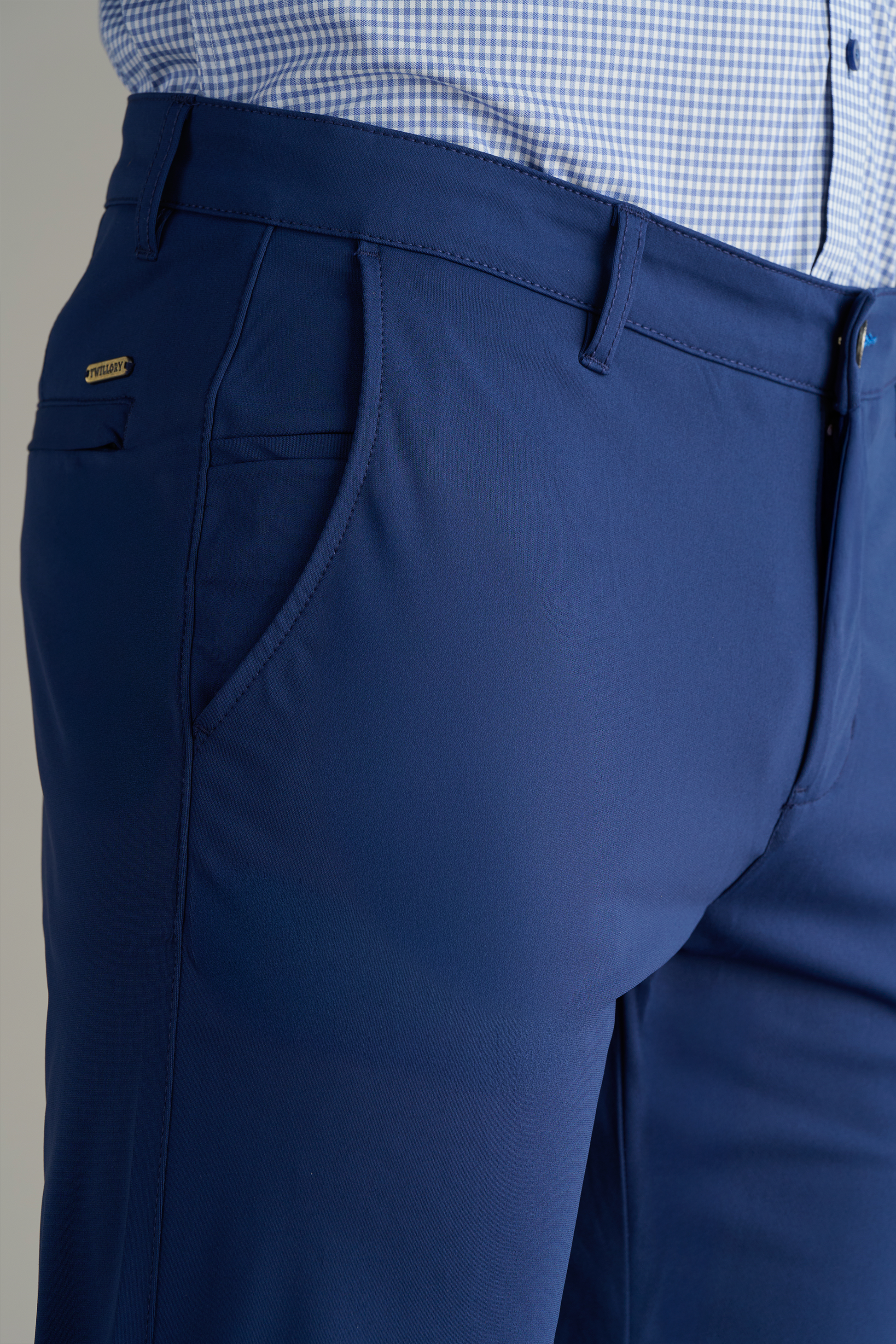 Close-up of a person wearing Twillory's Performance Pants in Cobalt Blue with a tucked-in light blue checkered shirt. Focus is on the upper thigh, waist, side pocket, belt loops, metal tag, and button details.
