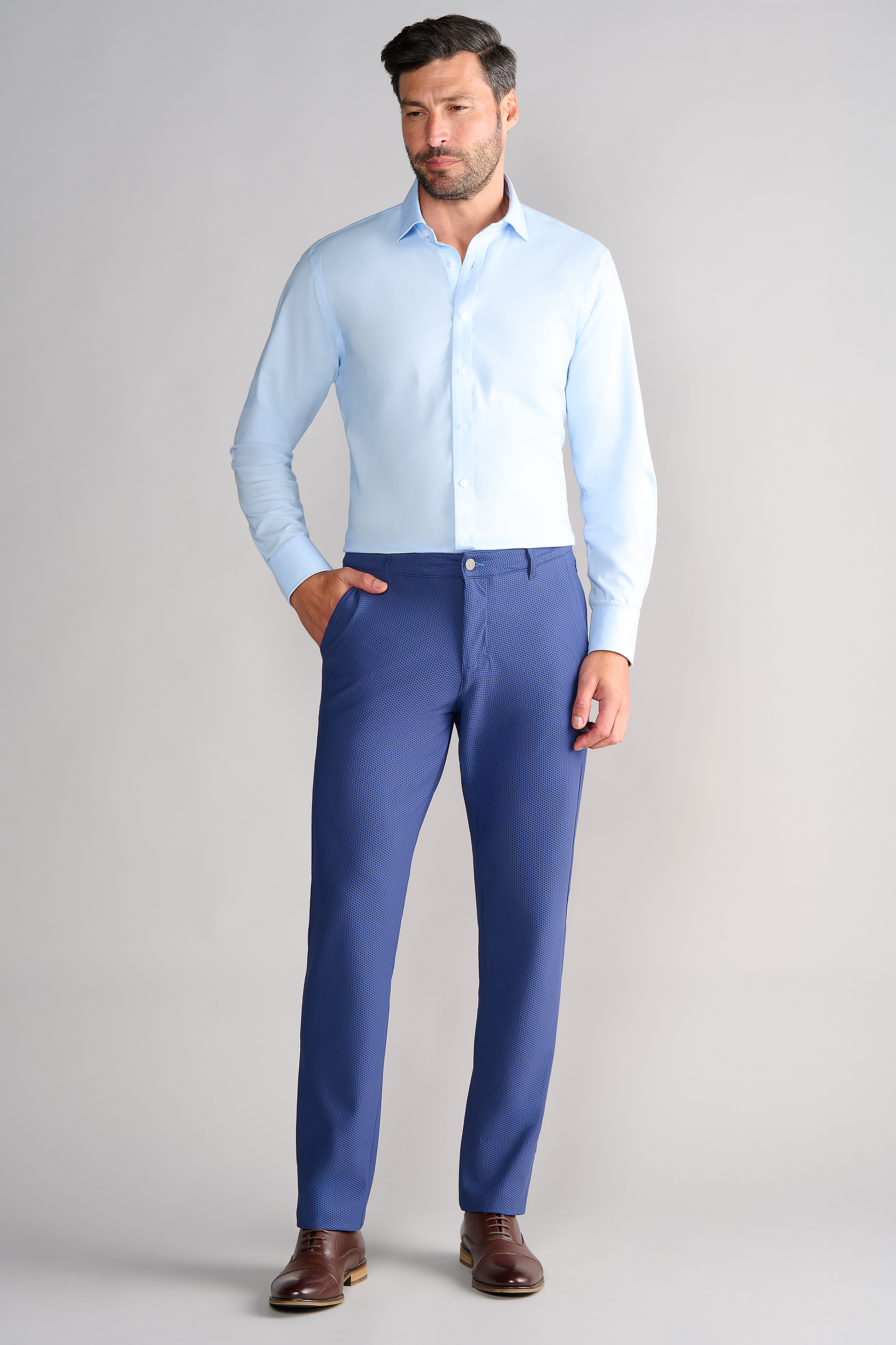 A man with short dark hair and a beard stands against a light gray background, wearing a light blue dress shirt and Twillory’s AIR Pants // Blue. He pairs them with brown dress shoes, one hand in his pocket as he looks thoughtfully to the side.