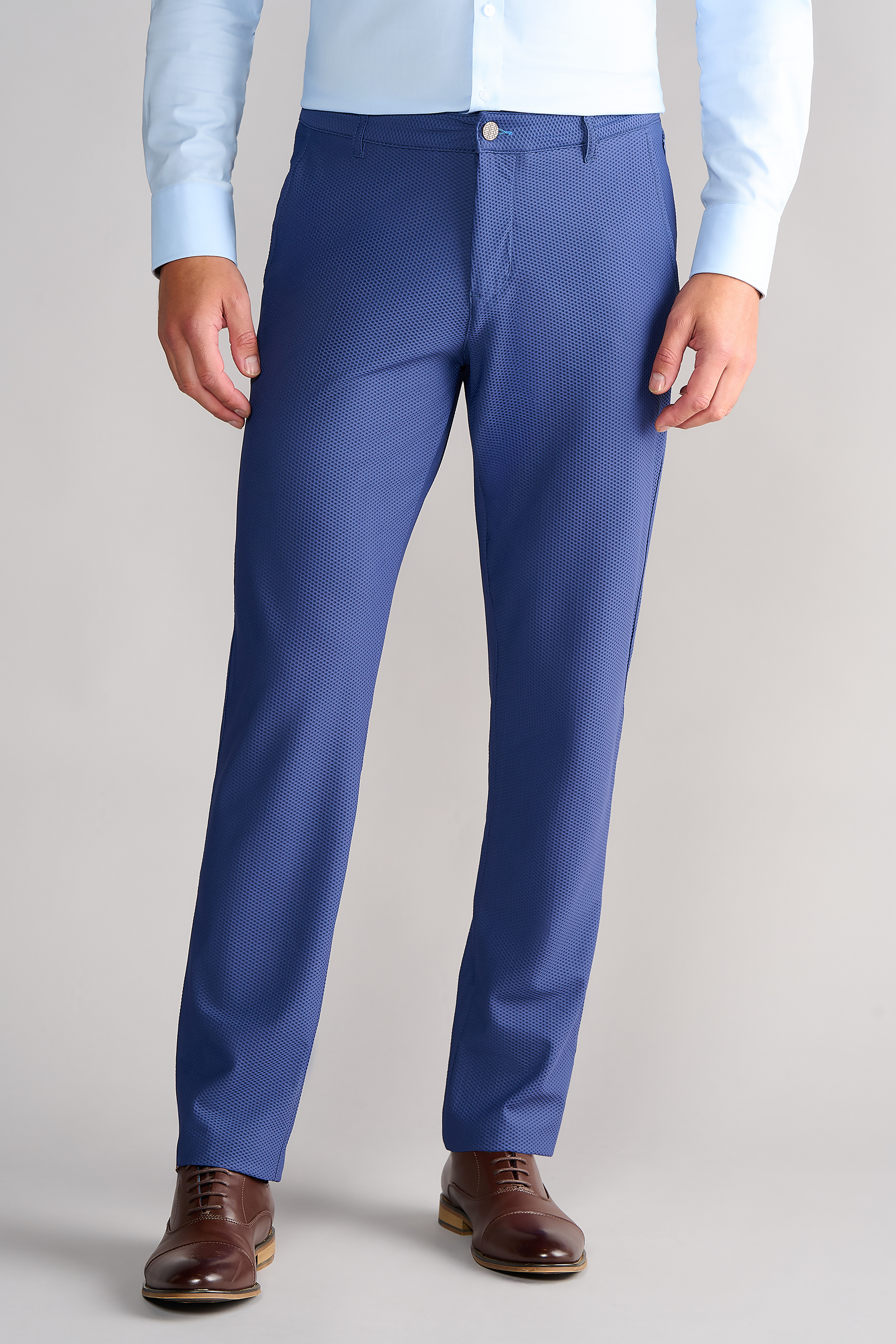 A person wears Twillory AIR Pants // Blue with a light blue dress shirt and brown Oxford shoes, standing against a plain light gray background. The photo is cropped at the shoulders, showing just the lower torso and legs.