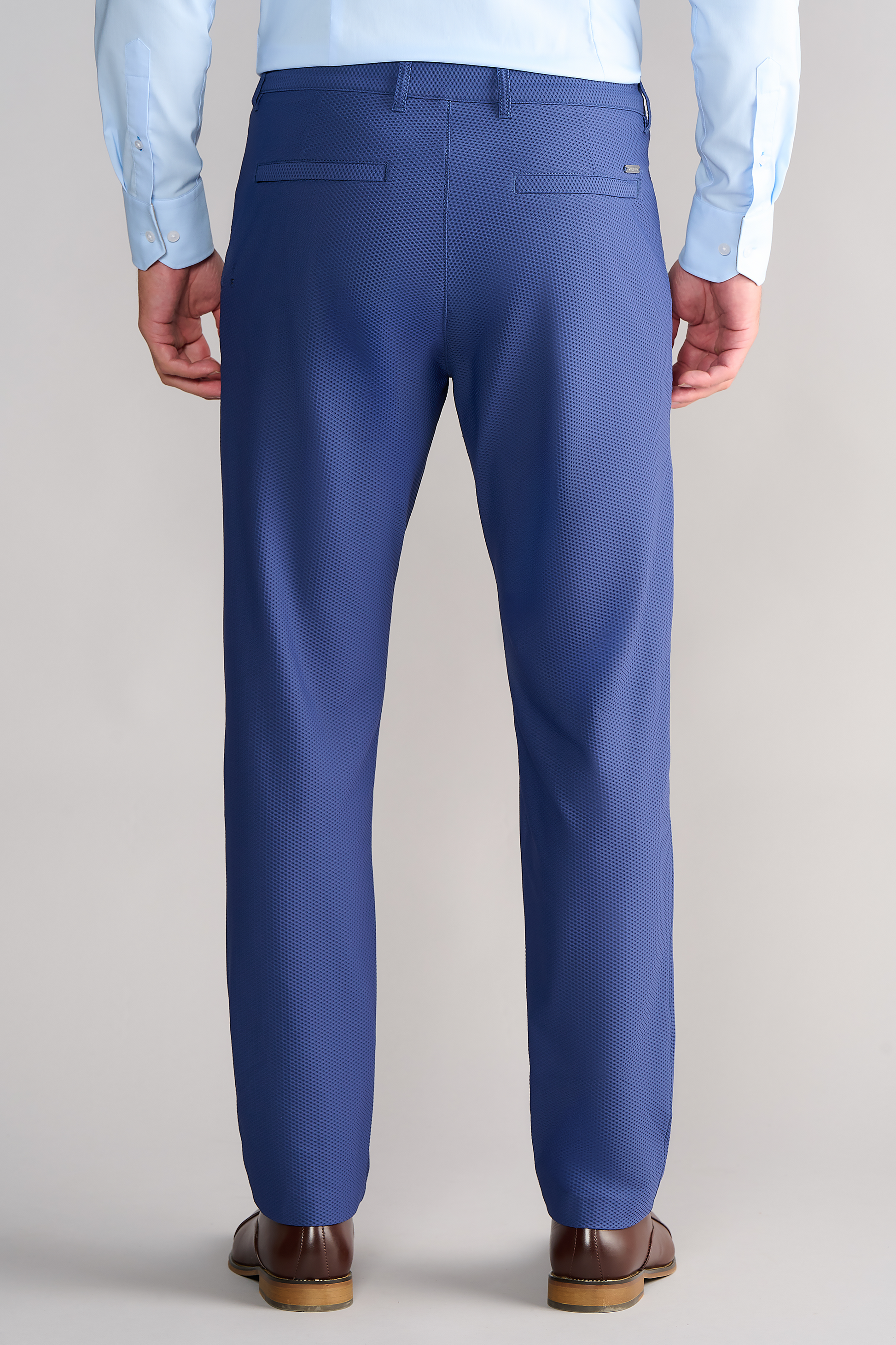 A person is shown from the back, wearing Twillory AIR Pants // Blue with 4-way stretch, welt pockets, and moisture-wicking textured fabric, paired with a light blue button-down shirt and brown dress shoes on a neutral background.