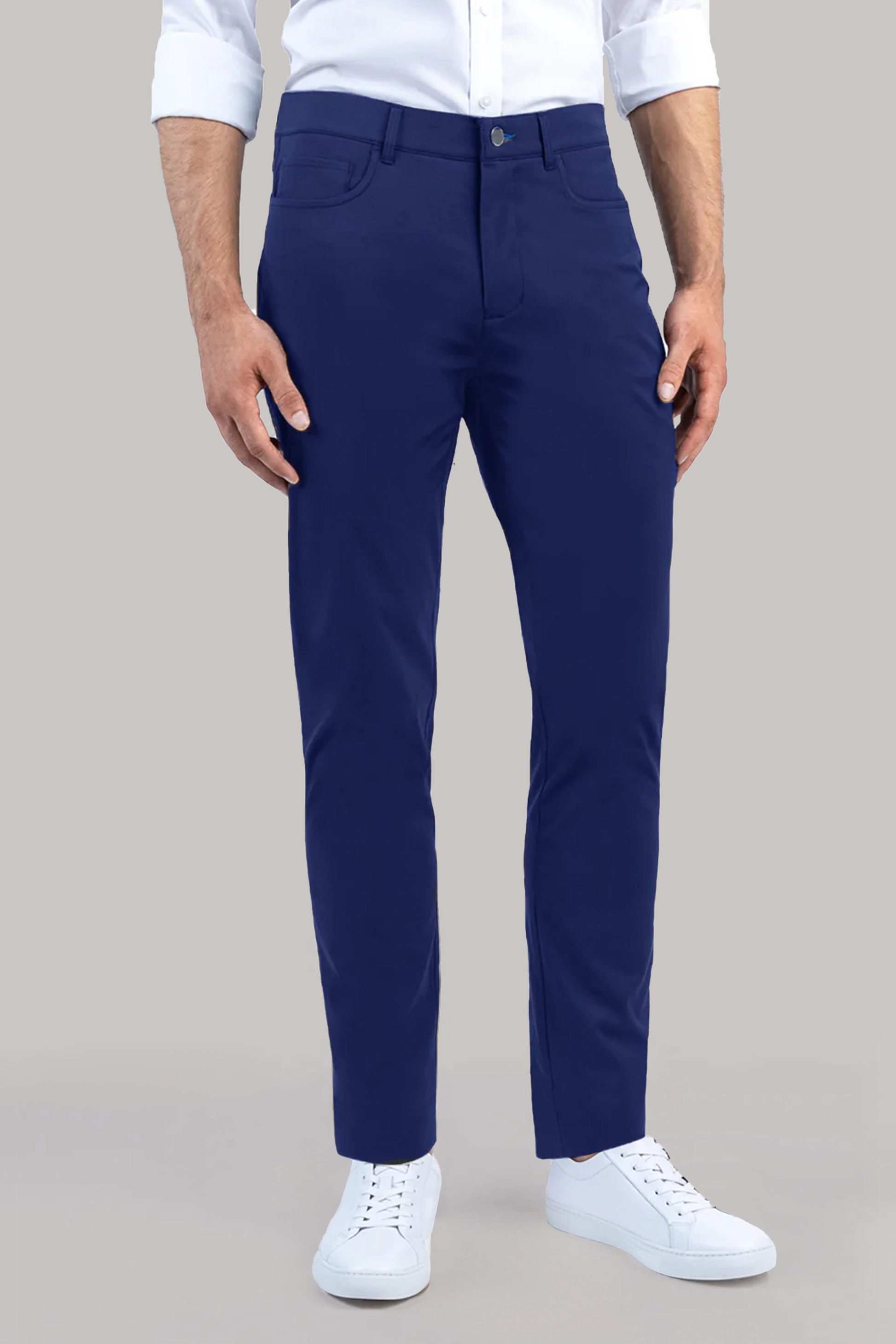 A man stands against a plain background wearing Twillory’s Performance Five Pocket pants in blue, a white 4-way stretch shirt, and white low-top sneakers. His hands rest at his sides, visible from shoulders to feet.
