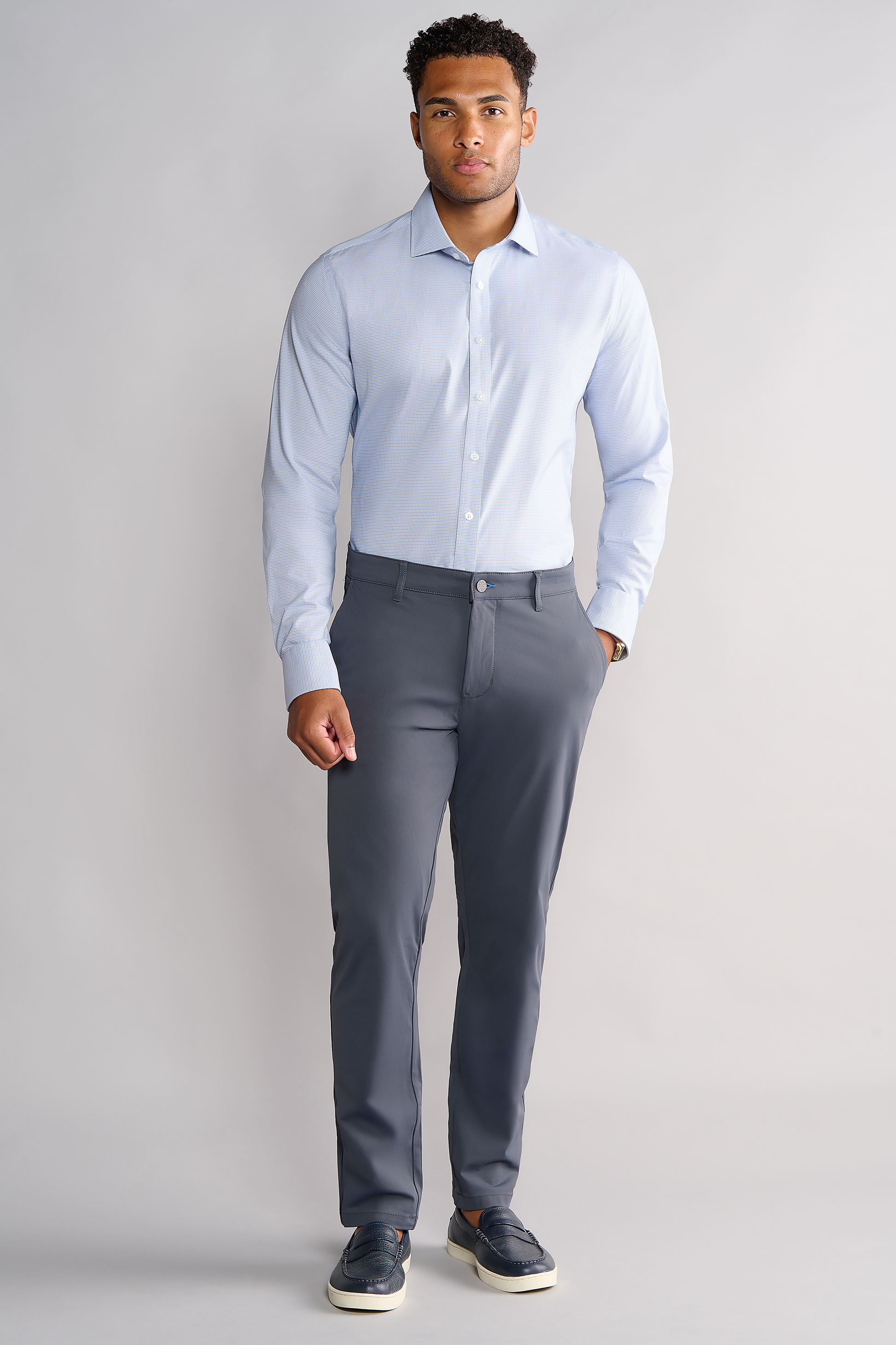 A man with short curly hair stands against a light grey background, wearing Twillory’s Leader Contrast // Blue Check shirt. He pairs it with grey dress pants and dark slip-on shoes, one hand by his side and the other in his pocket.