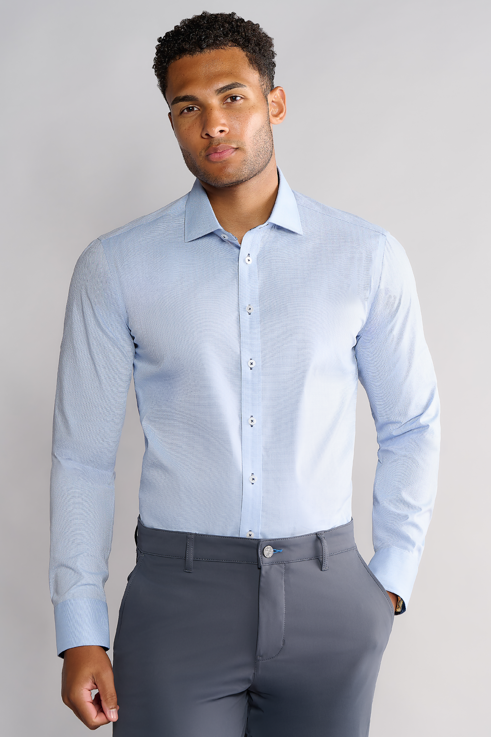 A man with short, curly hair and a neutral expression stands facing forward against a light gray background, wearing the Twillory Leader Contrast // Blue Check shirt with white buttons and dark gray dress pants. His left hand rests by his side.