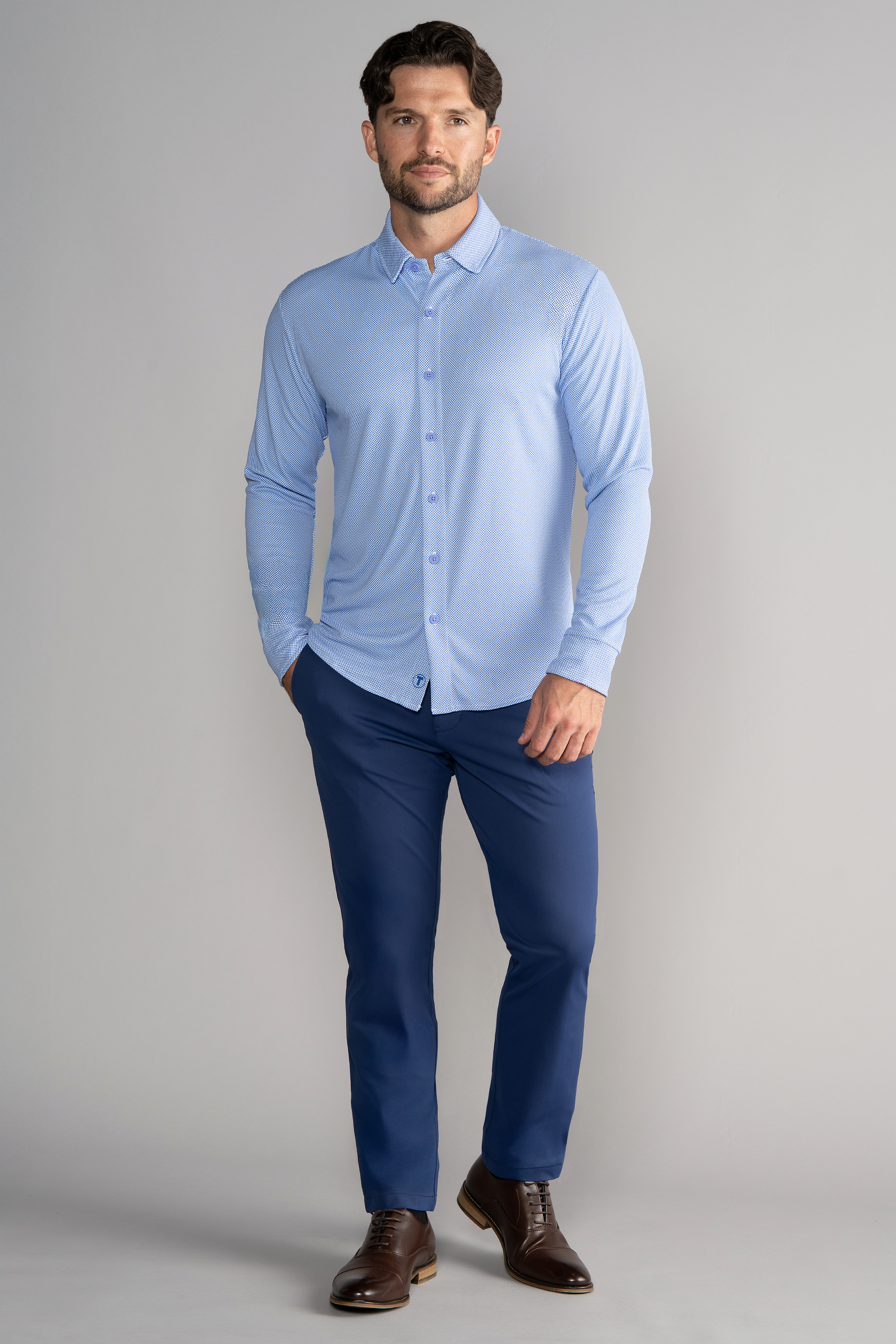 A man with short brown hair and a beard stands facing forward against a light gray background, wearing Twillory’s Long Sleeve Button Down Polo Dot in Blue Diamond, navy trousers, and brown leather dress shoes, with his left hand in his pocket.