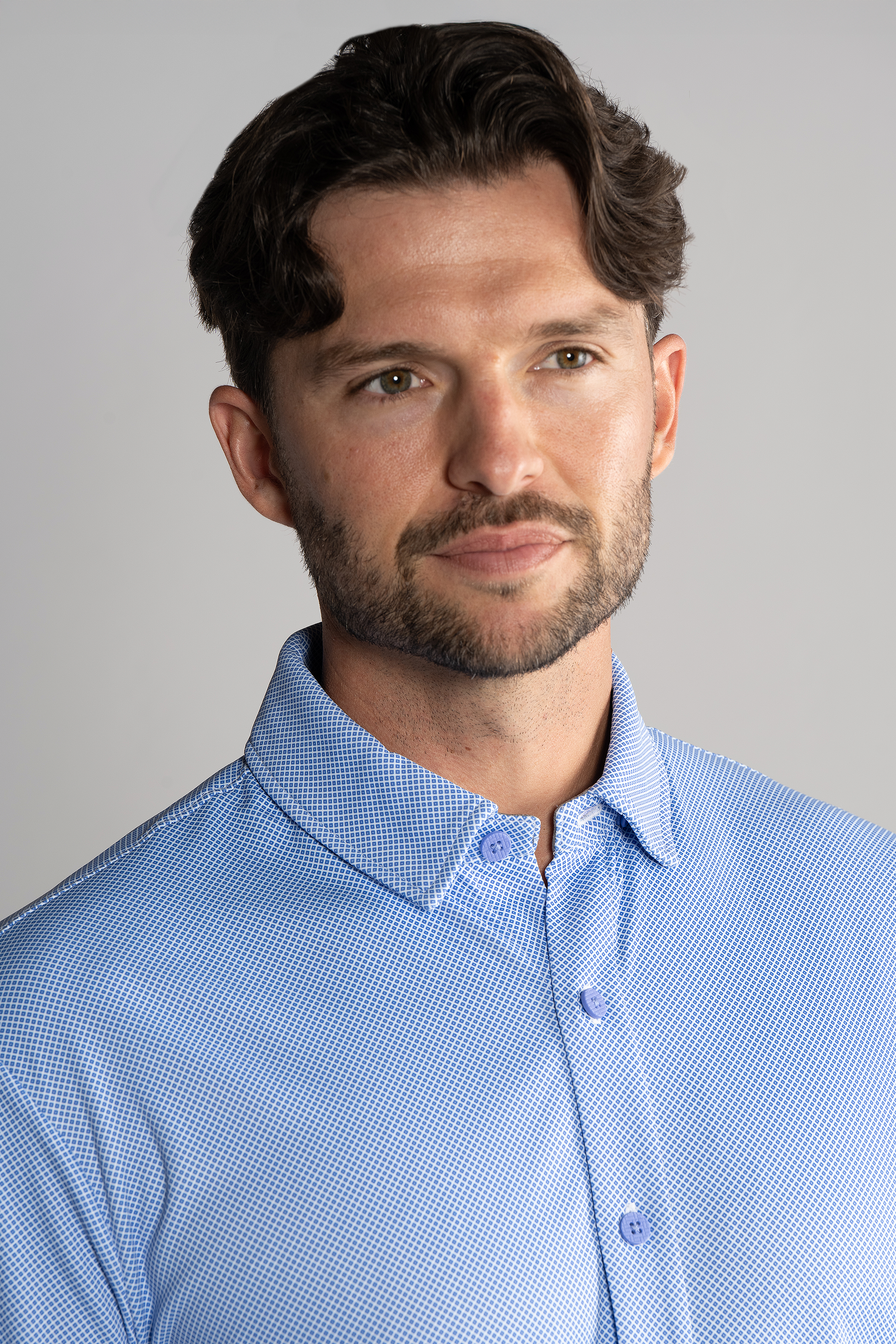A man with short brown hair and a trimmed beard wears the Twillory Long Sleeve Button Down Polo Dot in Blue Diamond, facing slightly right with a neutral expression against a plain light gray background.