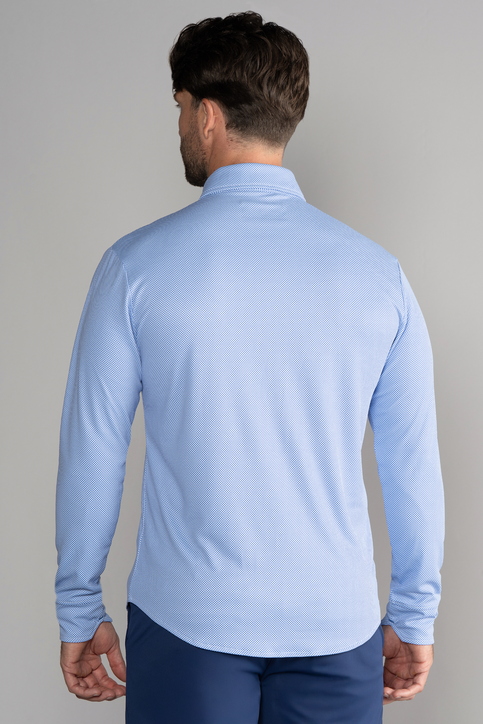 A man with short brown hair stands against a gray background, wearing the Twillory Long Sleeve Button Down Polo Dot // Blue Diamond—a moisture-wicking, light blue shirt with a subtle textured pattern and dark blue pants.