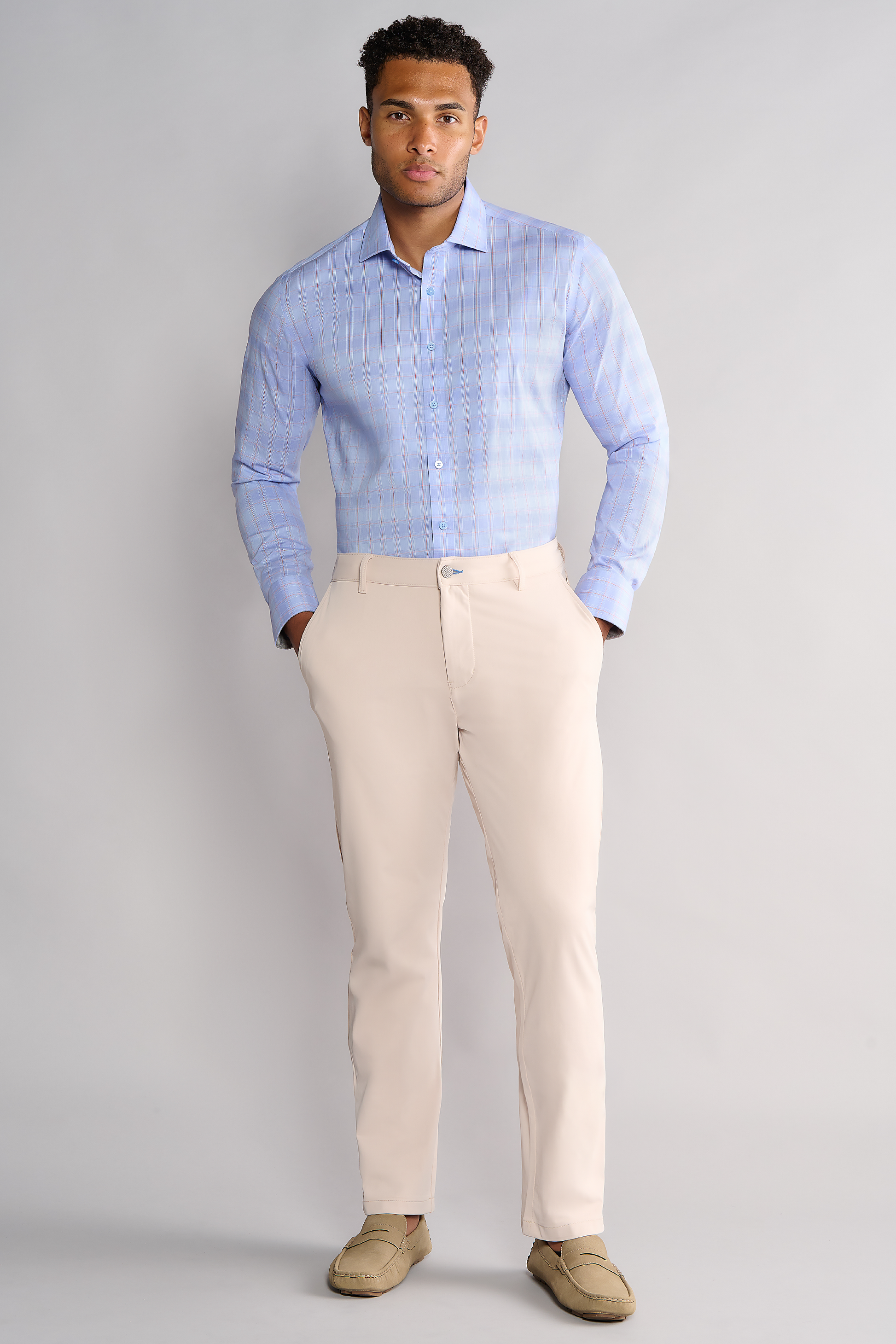 A man with short, curly hair and a neutral expression stands against a light gray background wearing Twillory's Leader Plaid // Blue Prince of Wales non-iron dress shirt, beige pants, and light tan loafers with his hands in his pockets.