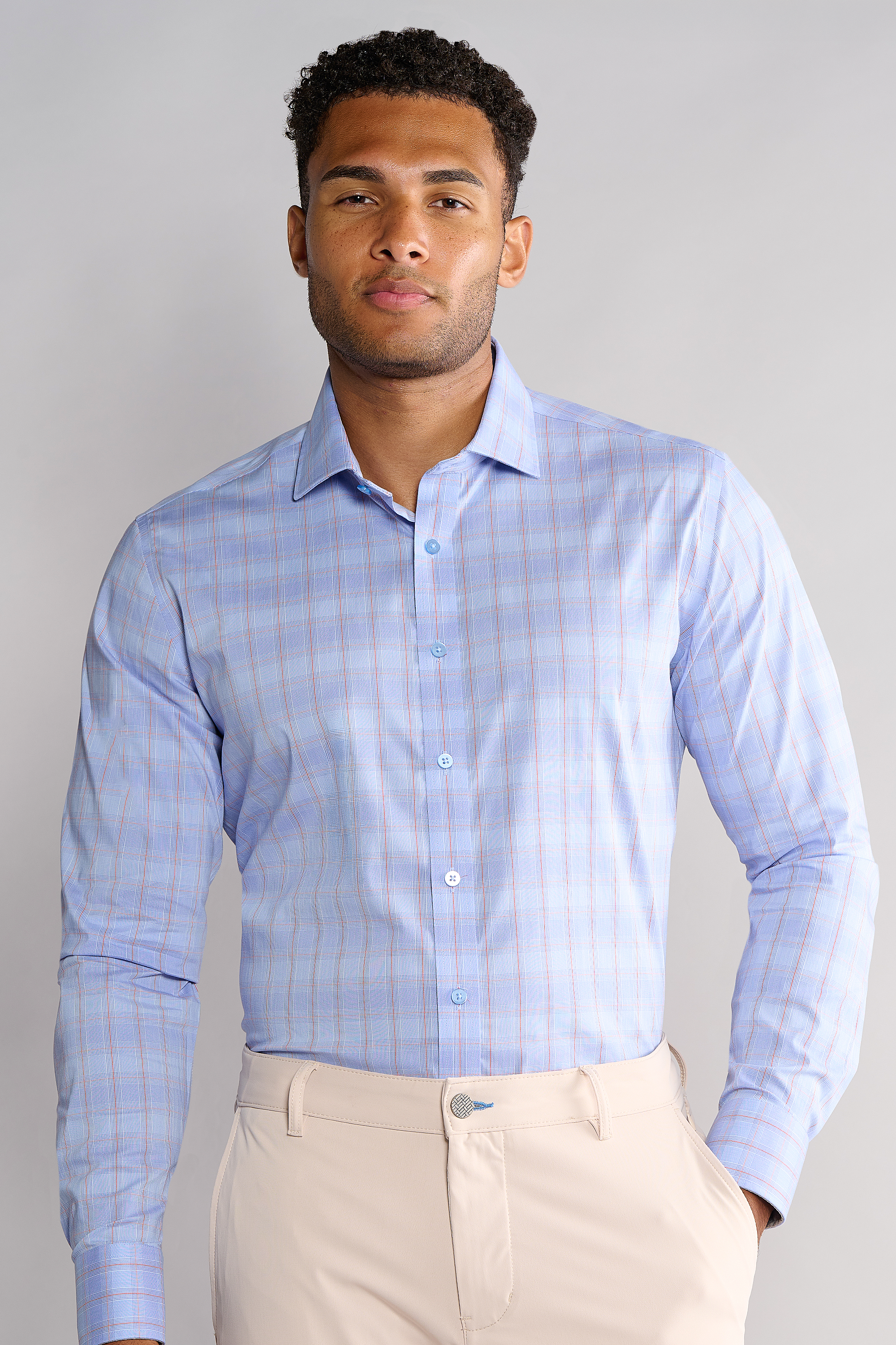 A man with short curly hair wears Twillory’s Leader Plaid // Blue Prince of Wales performance shirt—light blue with a subtle plaid. He stands confidently, one hand in light beige pants, against a plain light gray background.