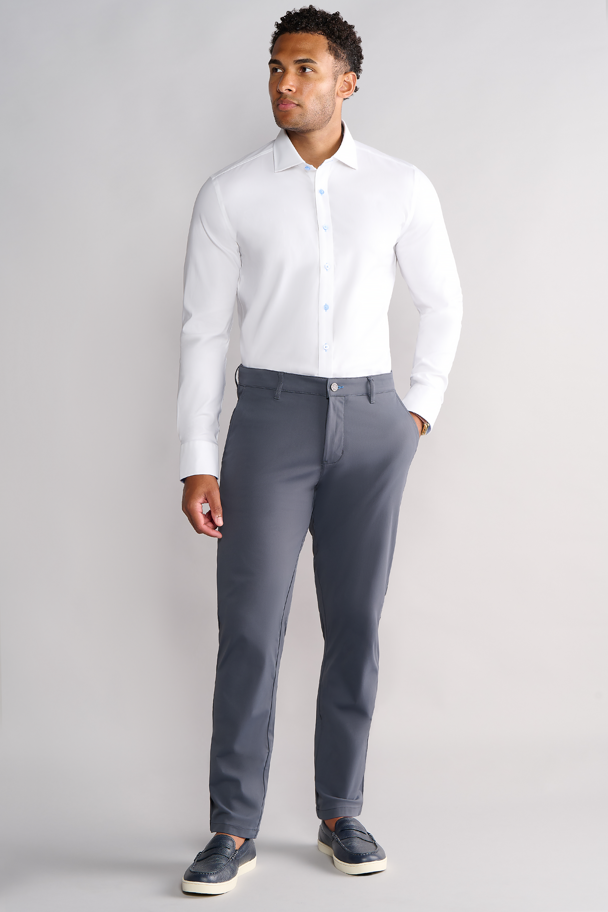 A man stands facing forward in Twillory's Leader Contrast // Blue Prince of Wales shirt against a plain background, paired with gray dress pants and dark slip-on shoes. His left hand is at his side and he gazes slightly right with a neutral look.